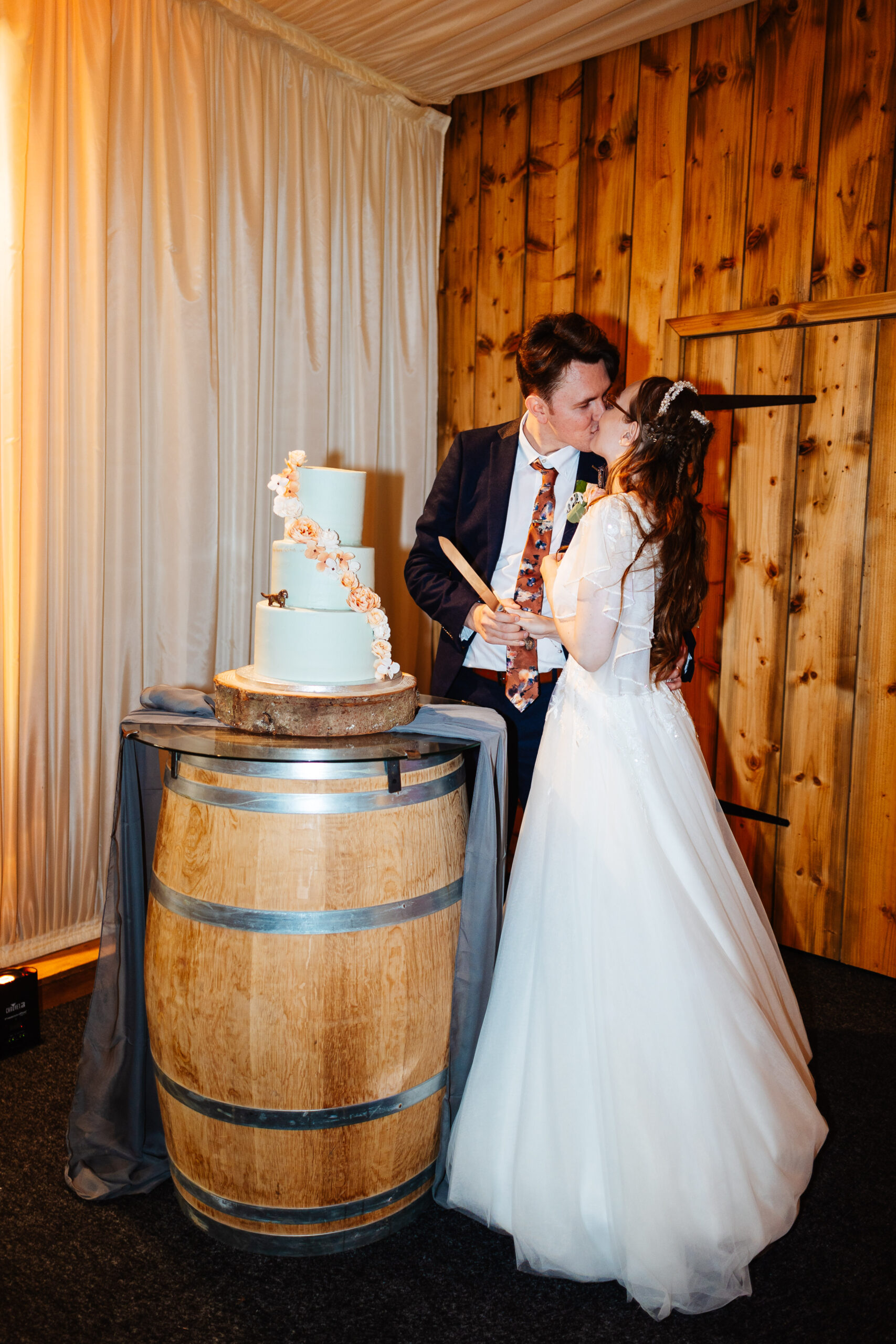 The bride and groom kissing. They are cutting the cake. The cake is a three tiered cake with little iced orange flowers down the tiers. There is a little toy spaniel on the side of the cake too.