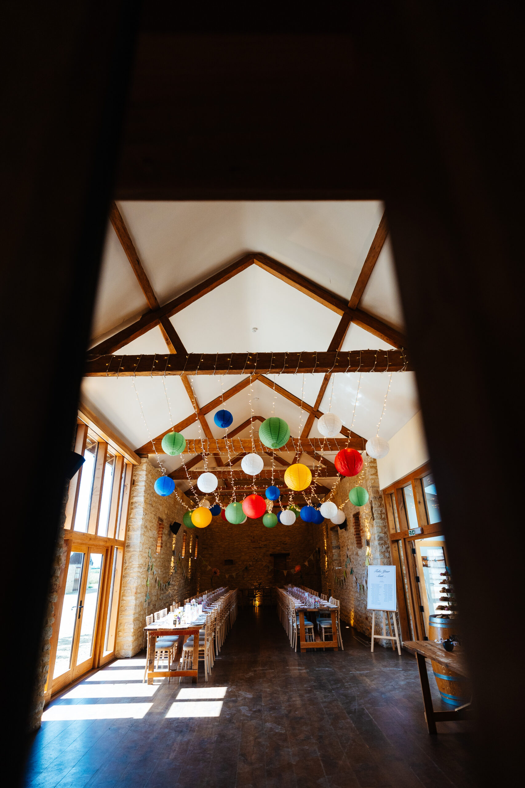 A large brick barn. There are long tables down the length of the hall and it has a dark brown wooden floor. There are bright coloured lanterns hanging from the wooden beams.