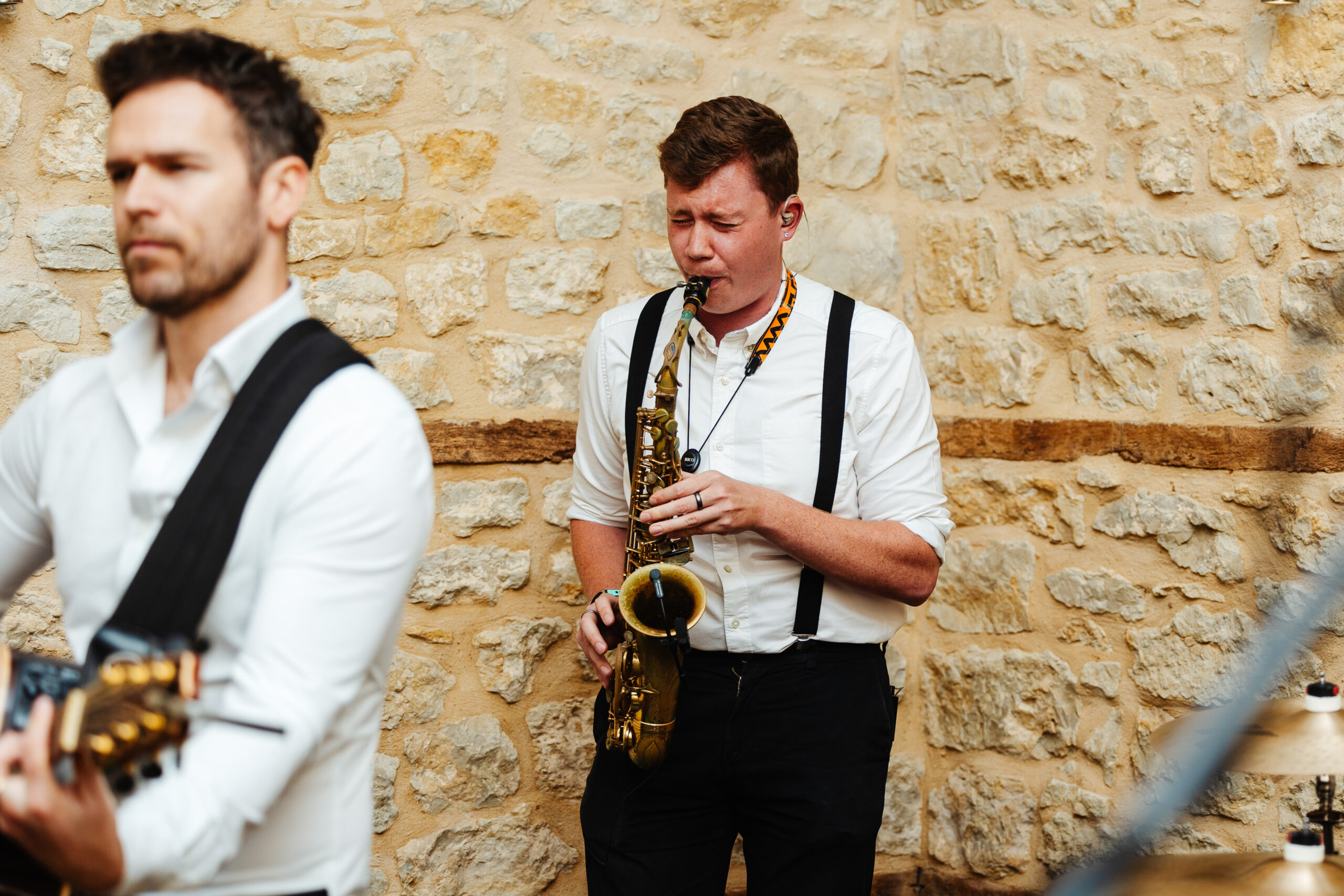 A photo of a saxophonist who is playing the saxphone. He is in a white shirt with black braces. His top button is undone.