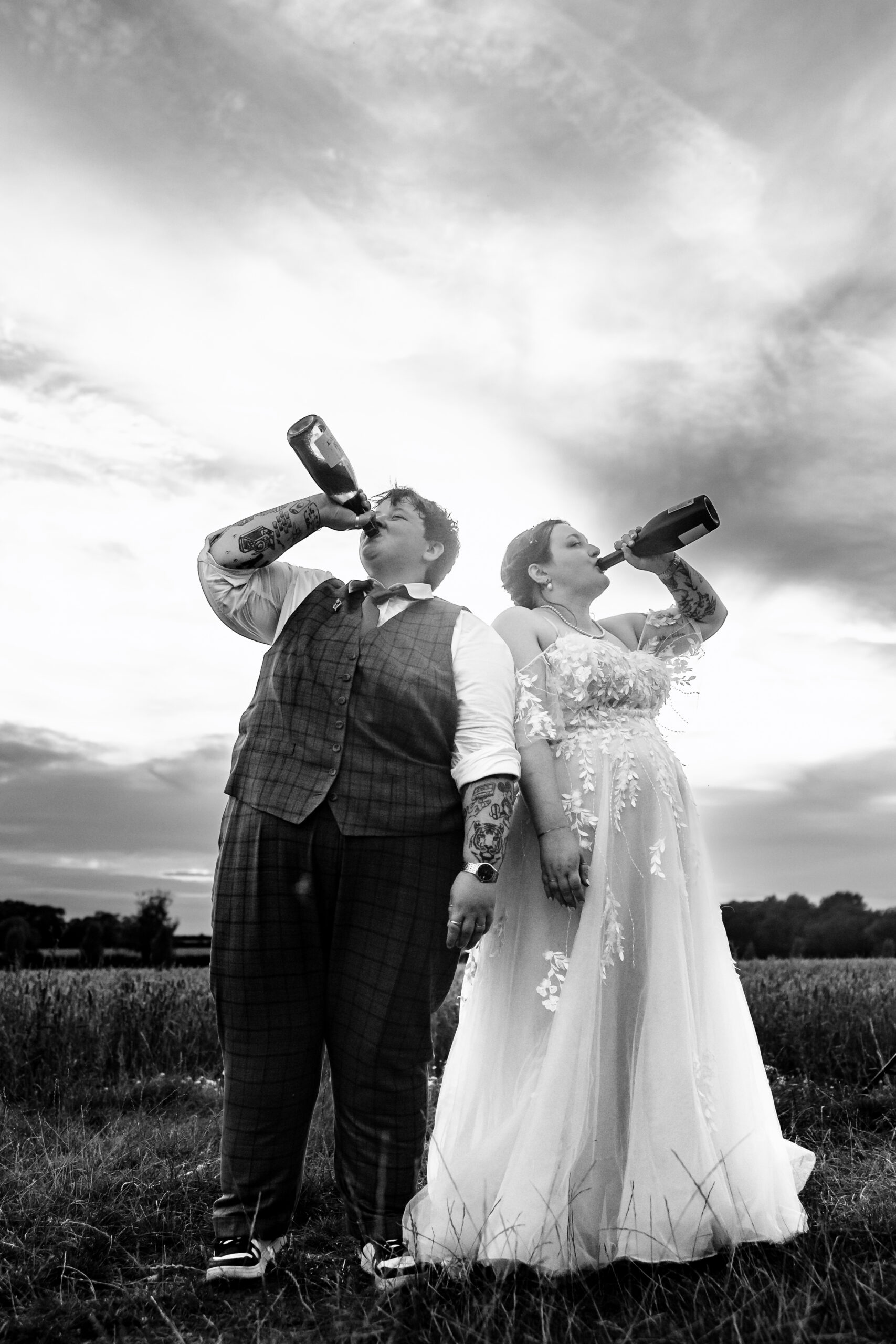 A black and white image of the couple in a corn field. They are both drinking a bottle of prosecco each and smiling.