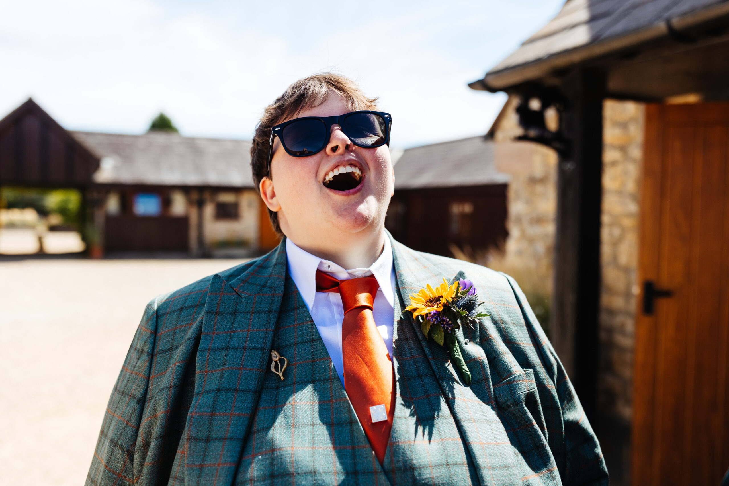 The bride in her suit. Her suit is turquoise with grey and orange stripes and she is wearing a burnt orange tie. She has sunglasses on and is laughing.