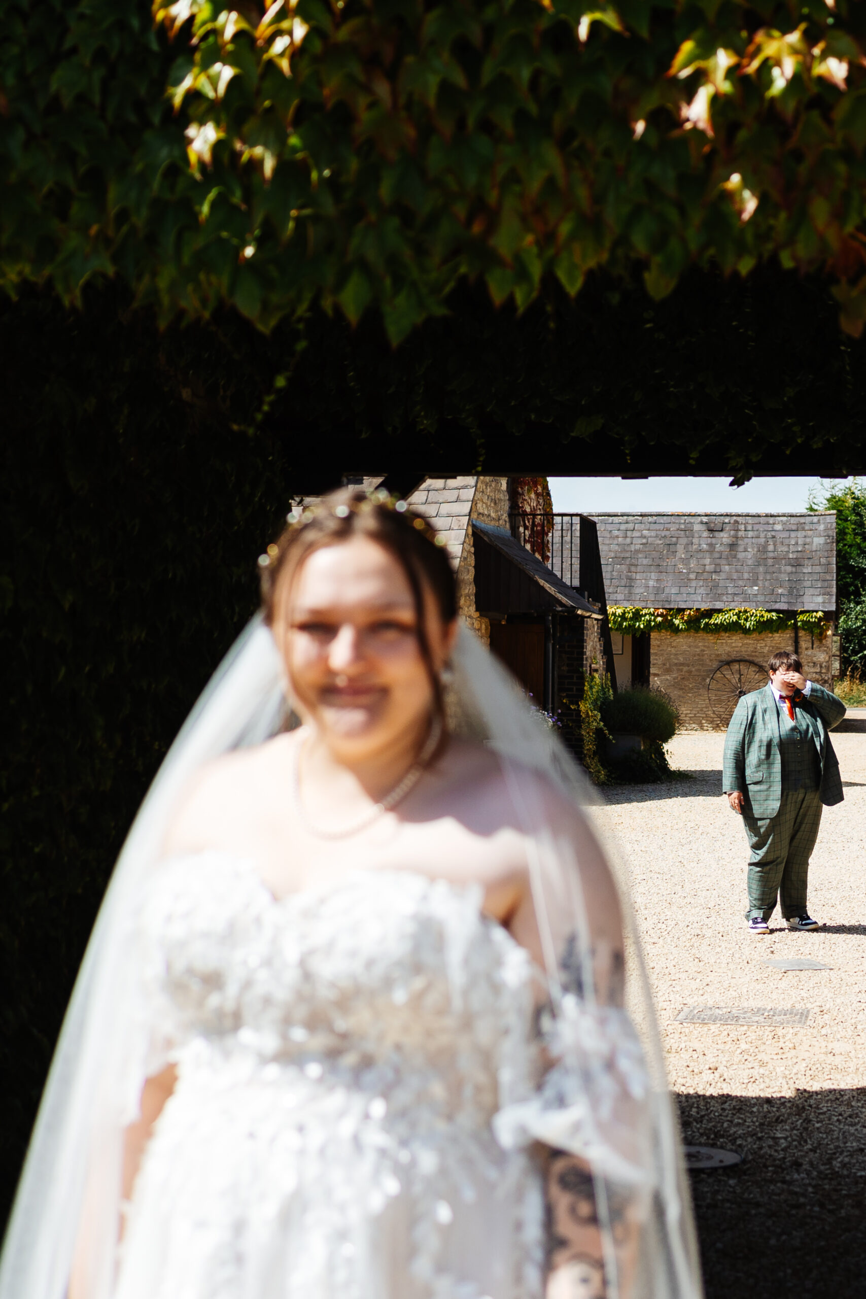 A photo of the two brides outside. One of the brides in her dress is out of focus. You can see the other bride behind her in focus and she has her hands over her eyes.