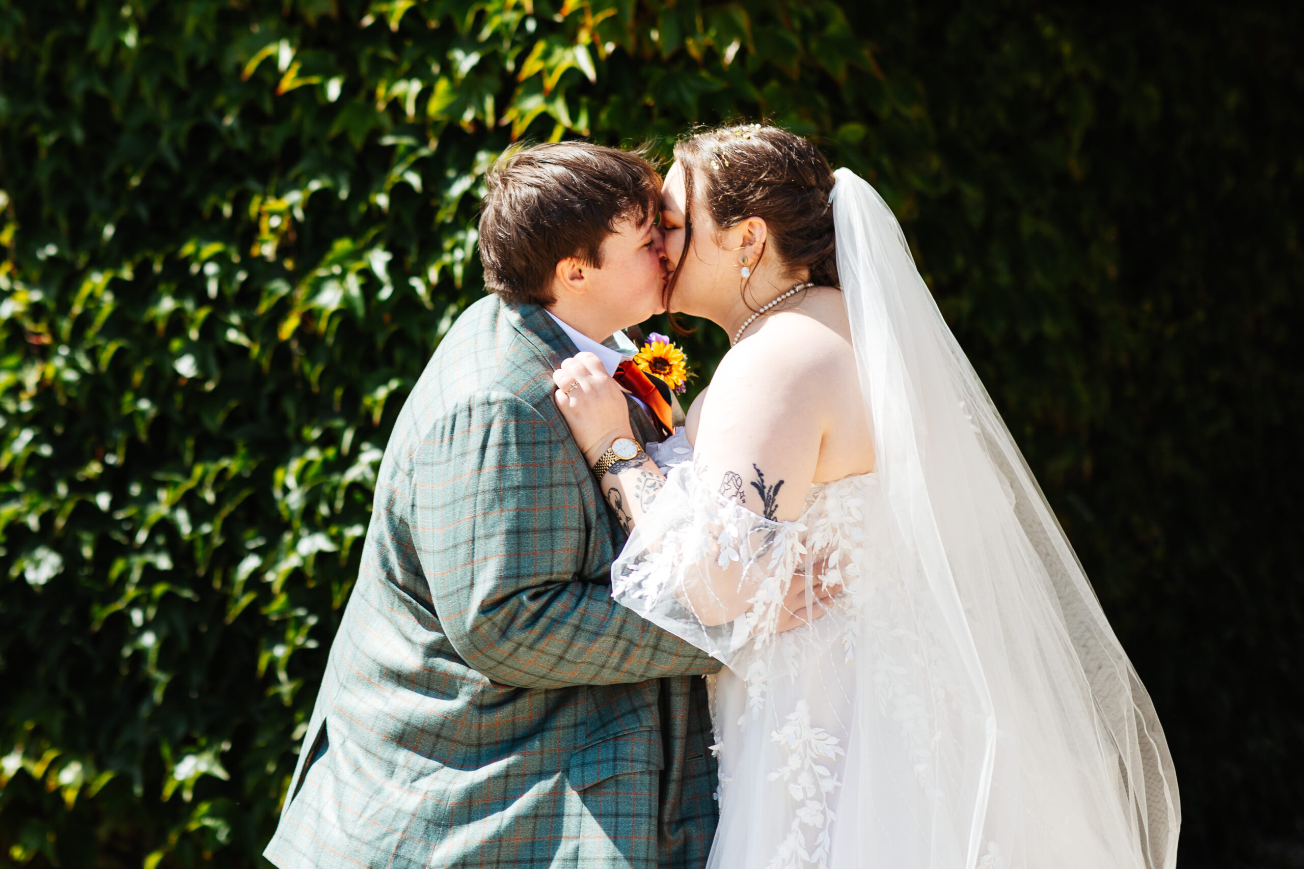 The brides kissing. They are stood behind a backdrop of luscious, green plants.