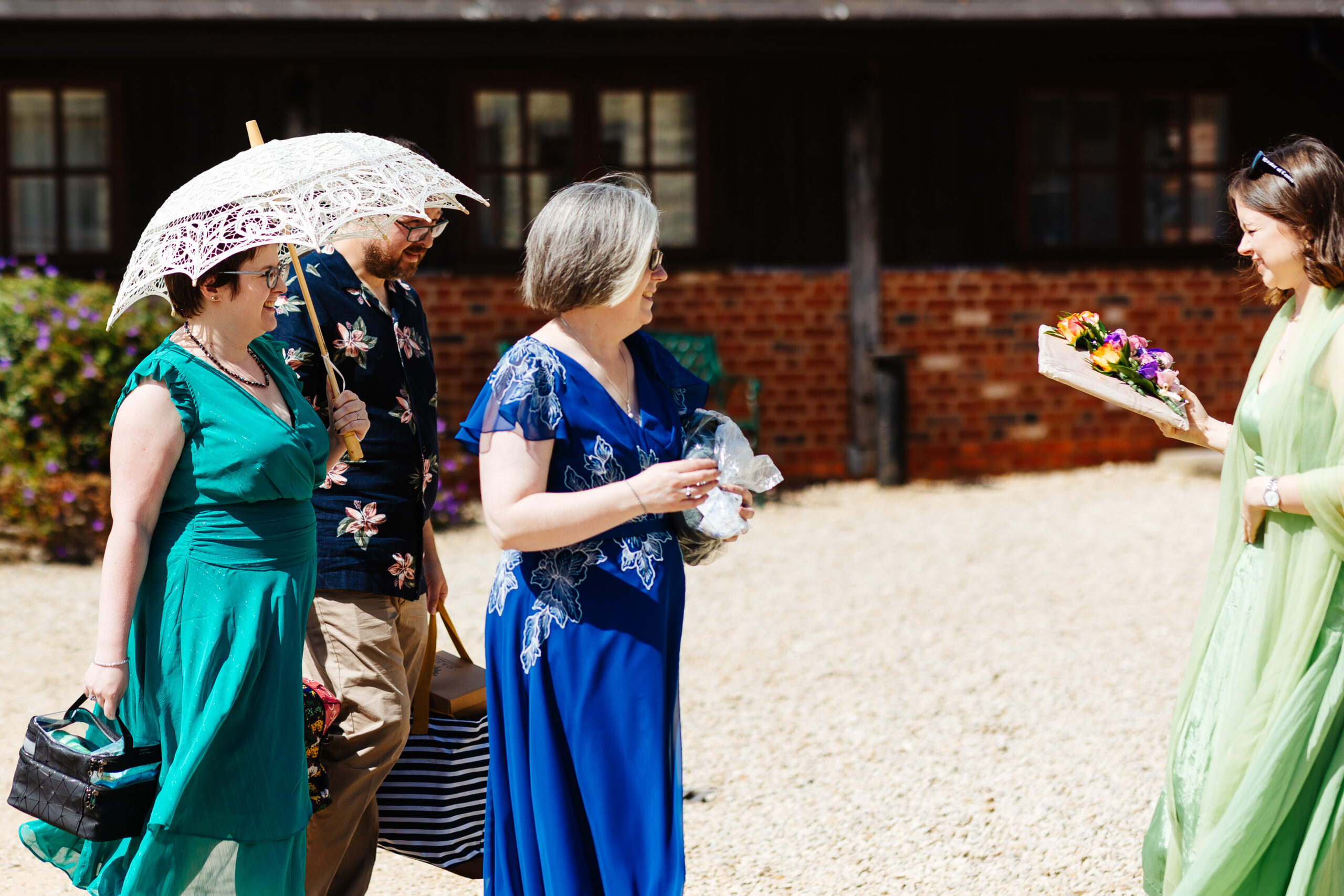 A photo of guests arriving. They are wearing bright colours and one of the guests is holding a white, lace umbrella. It is very sunny outside.