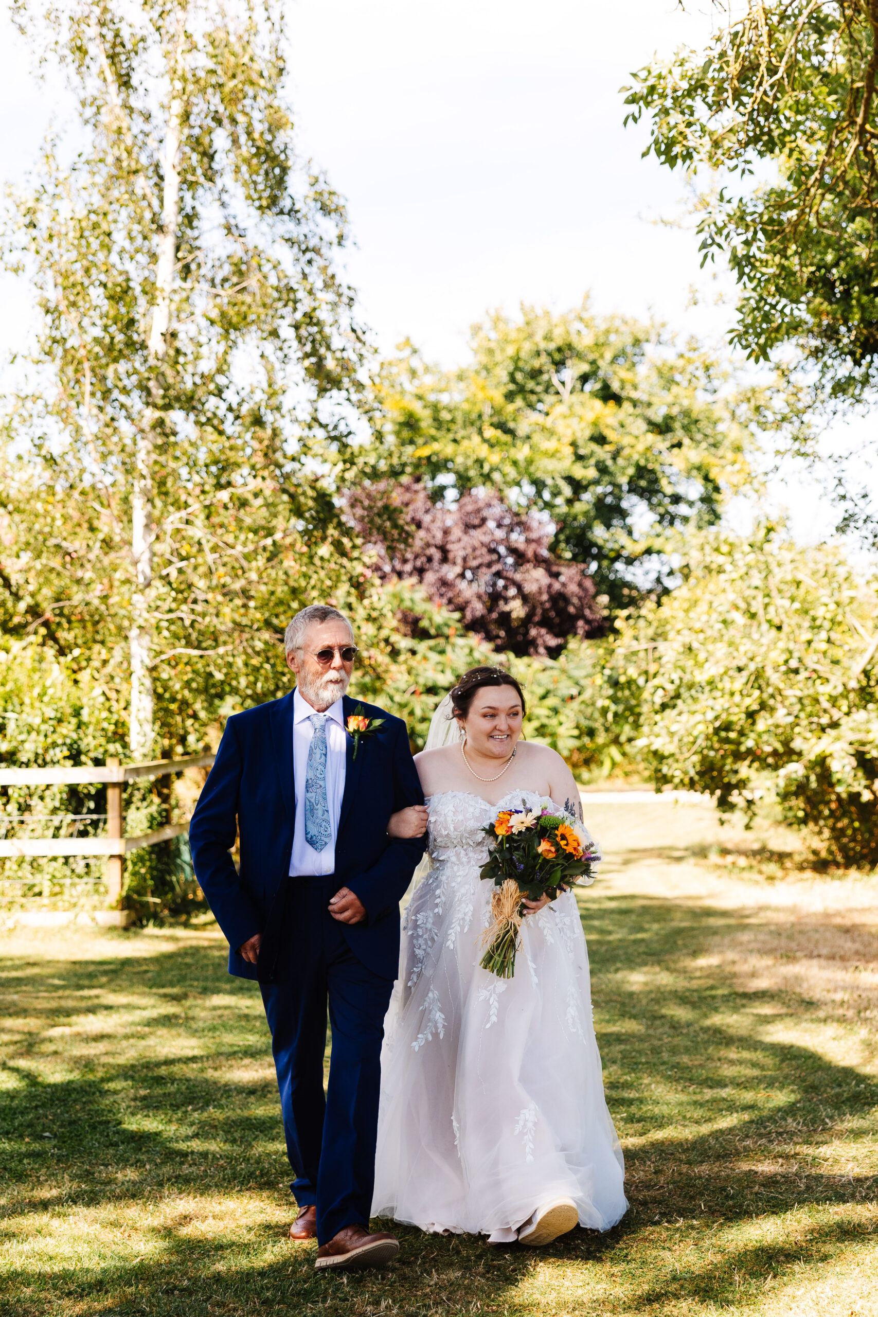 The bride in her dress holding her bouquet and walking down the aisle. She is arm in arm with her dad. 