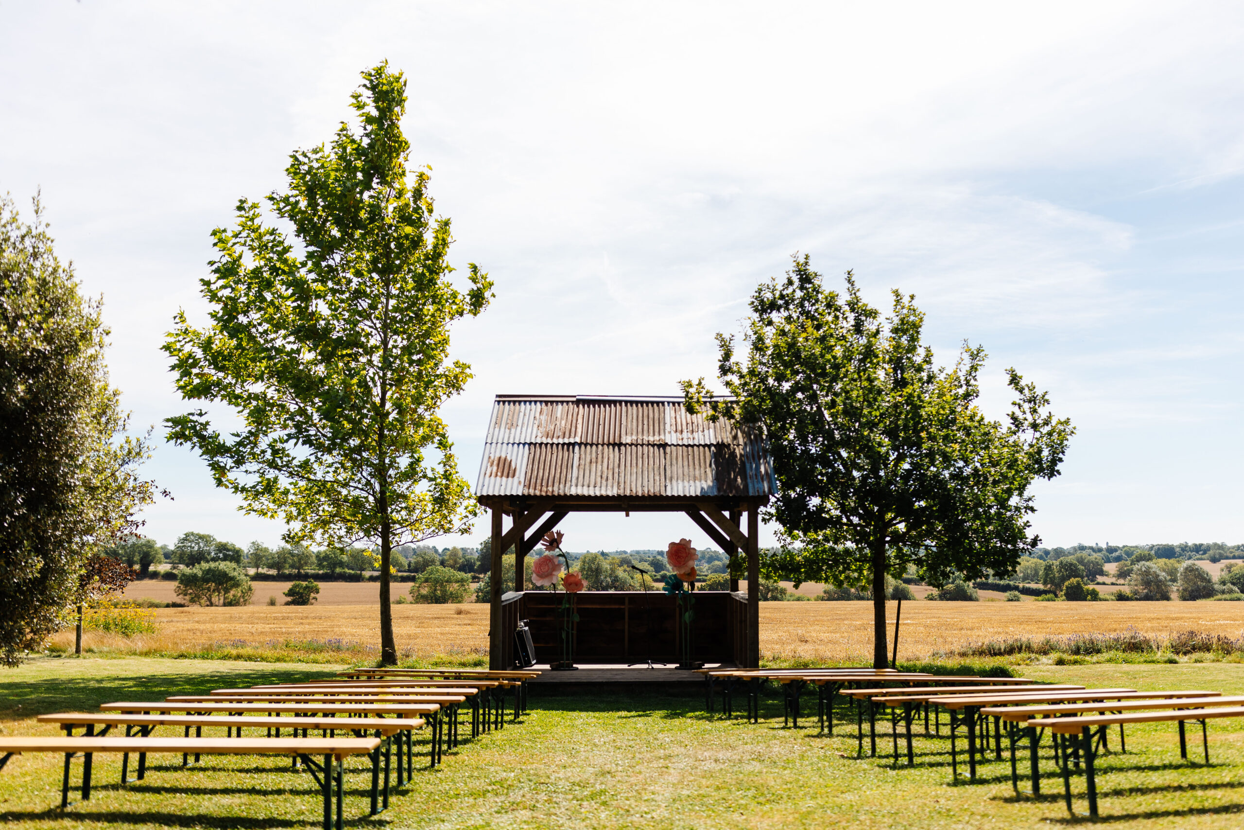 A picture of the outdoor setup for where the wedding will take place. There are wooden benches either side of the aisle and you can see a corrugated iron structure at the bottom wit very large, paper flowers in.