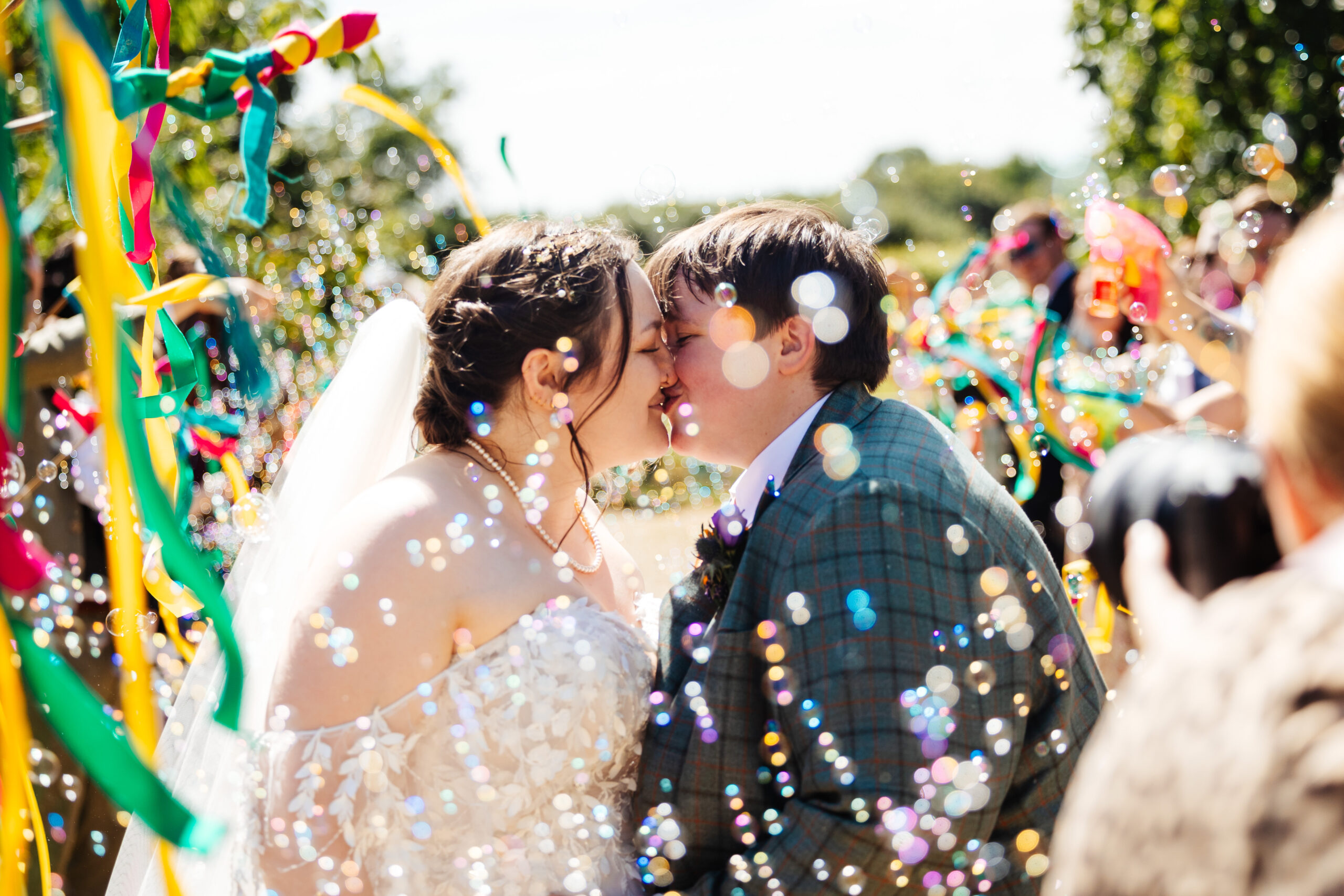 The brides kissing. You can see their guests in the background and they are carrying colourful ribbons. There are bubbles everywhere.