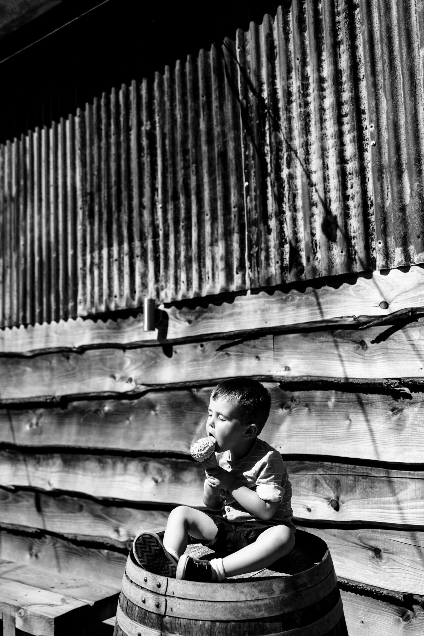 A black and white image of a little boy sitting on top of a wooden barrel. He is in a shirt and shorts and he is licking an ice cream.