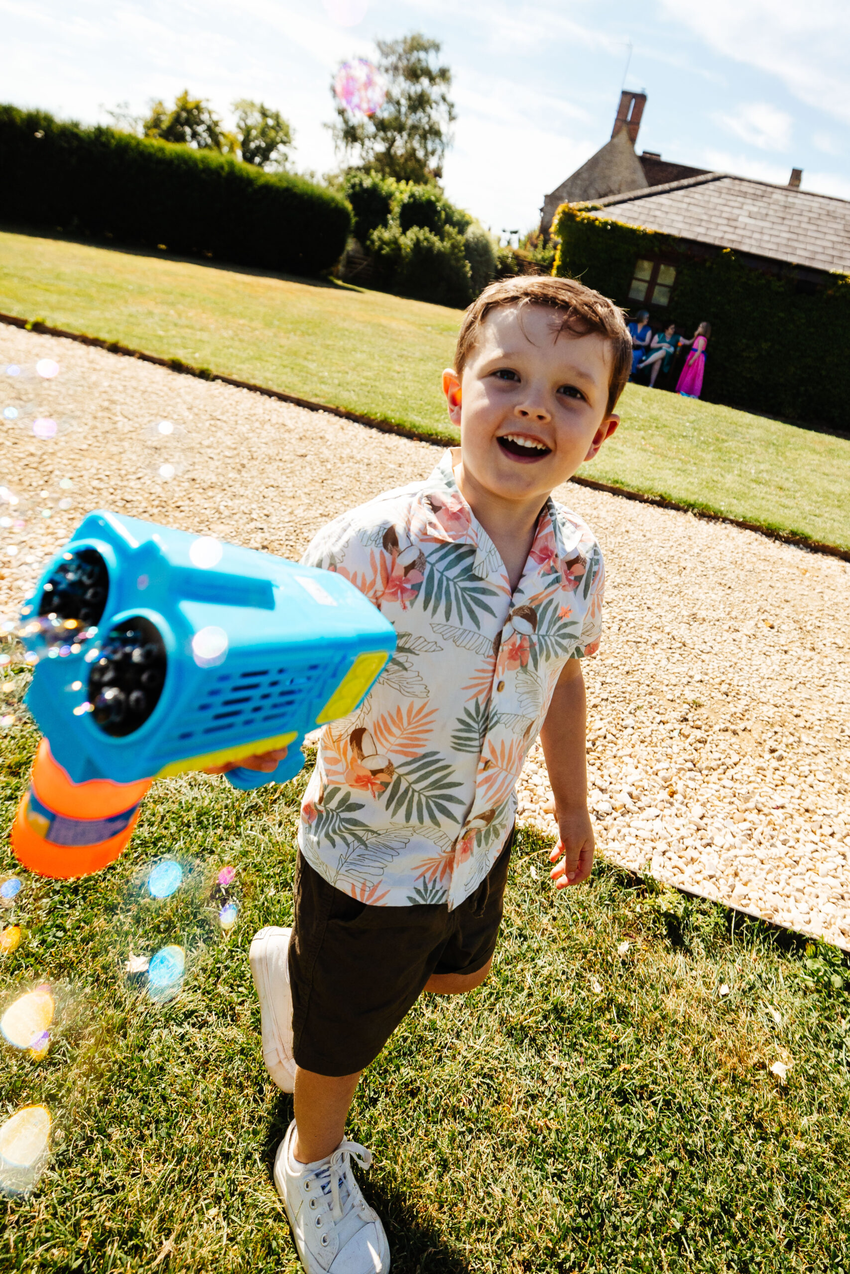 A little boy playing outside with a bubble gun. He is looking at the camera smiling and there are bubbles around him.