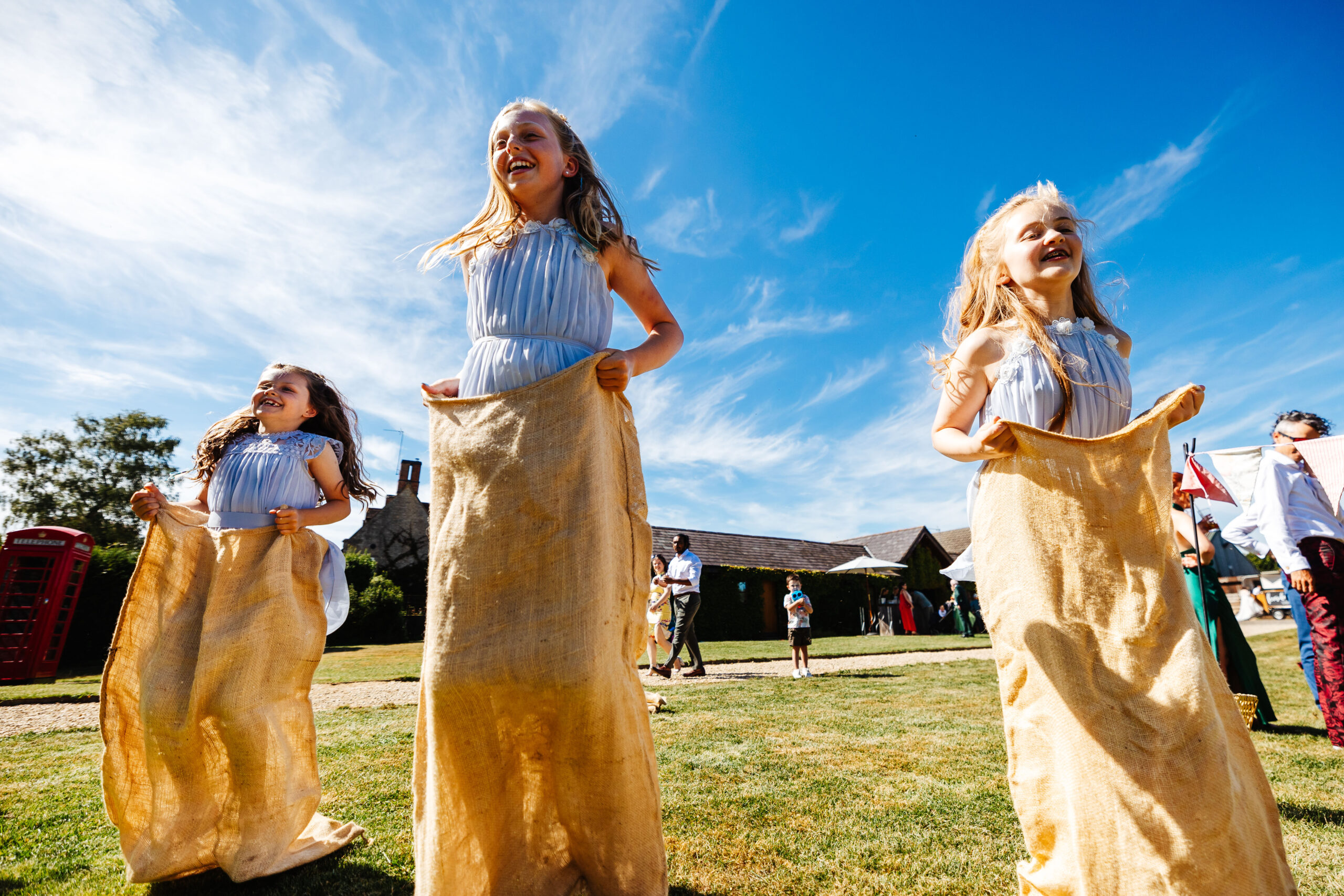 Three little girls in pale blue dresses. They are standing in hessian sacks having a race outside. The sky is bright blue.