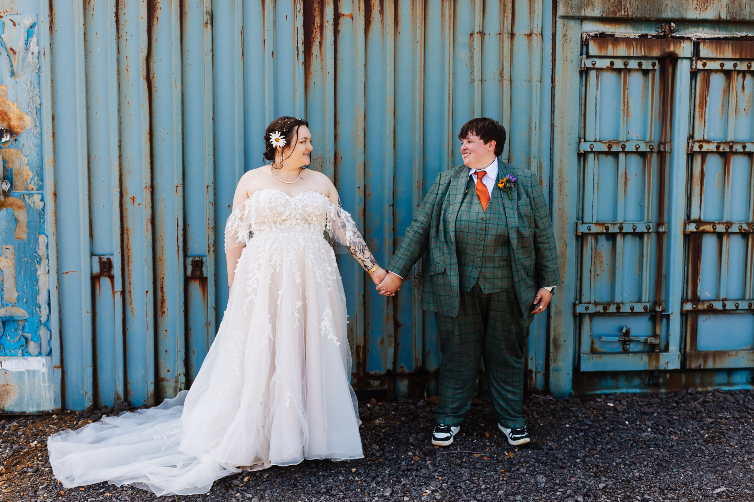 The brides in front of a corrugated iron, rustic container. They are holding hands and looking at one another smiling.