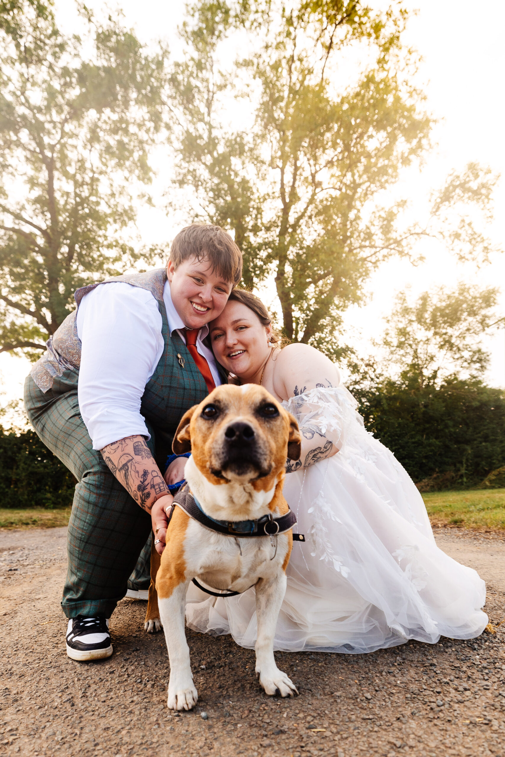 The brides outside and their dog. They are all looking at the camera and smiling and the dog is looking at the camera too.