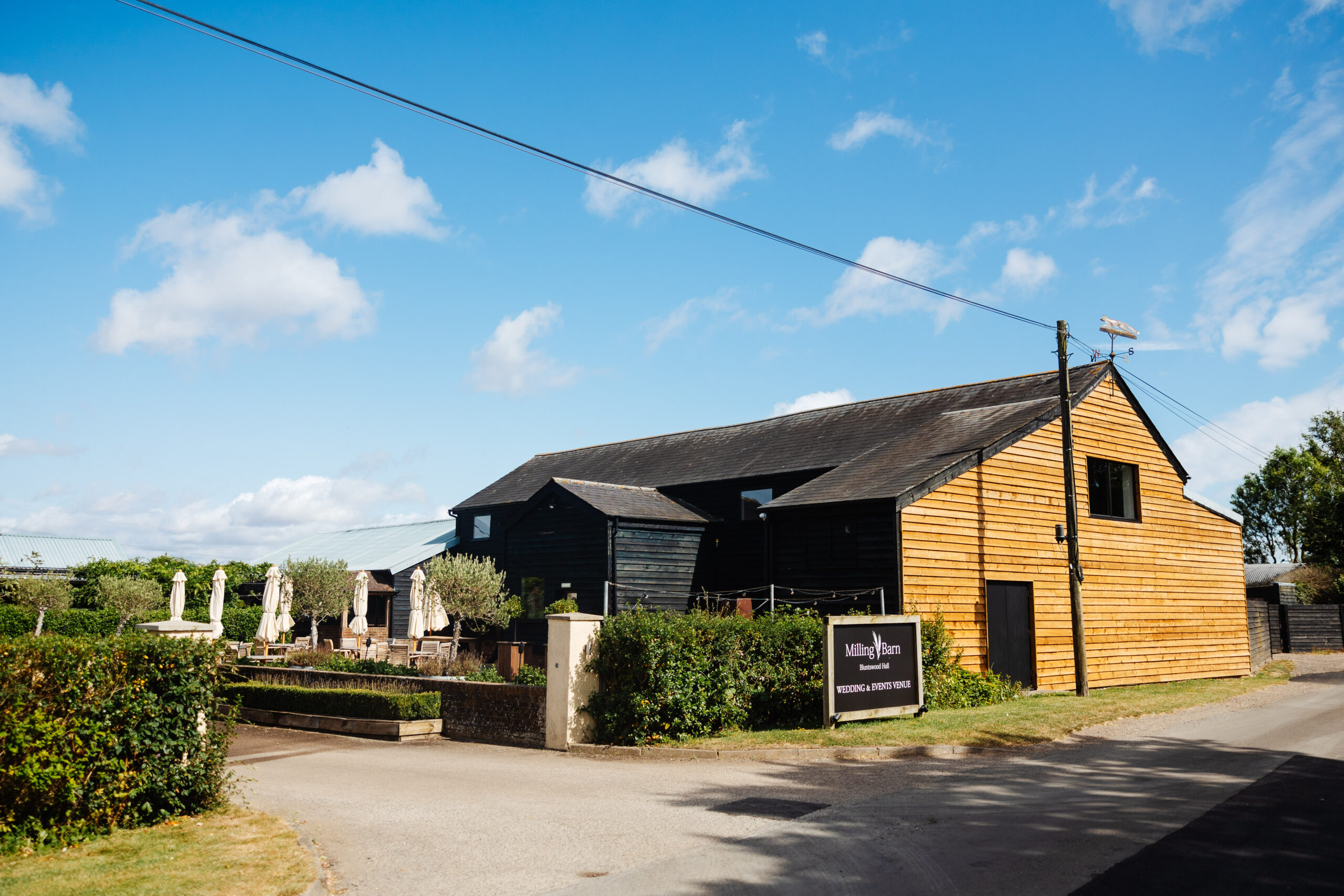 A photo of the venue - The Milling Barn. There is a wooden shed with Milling Barn signage on and the sky is bright blue with minimal, wispy clouds