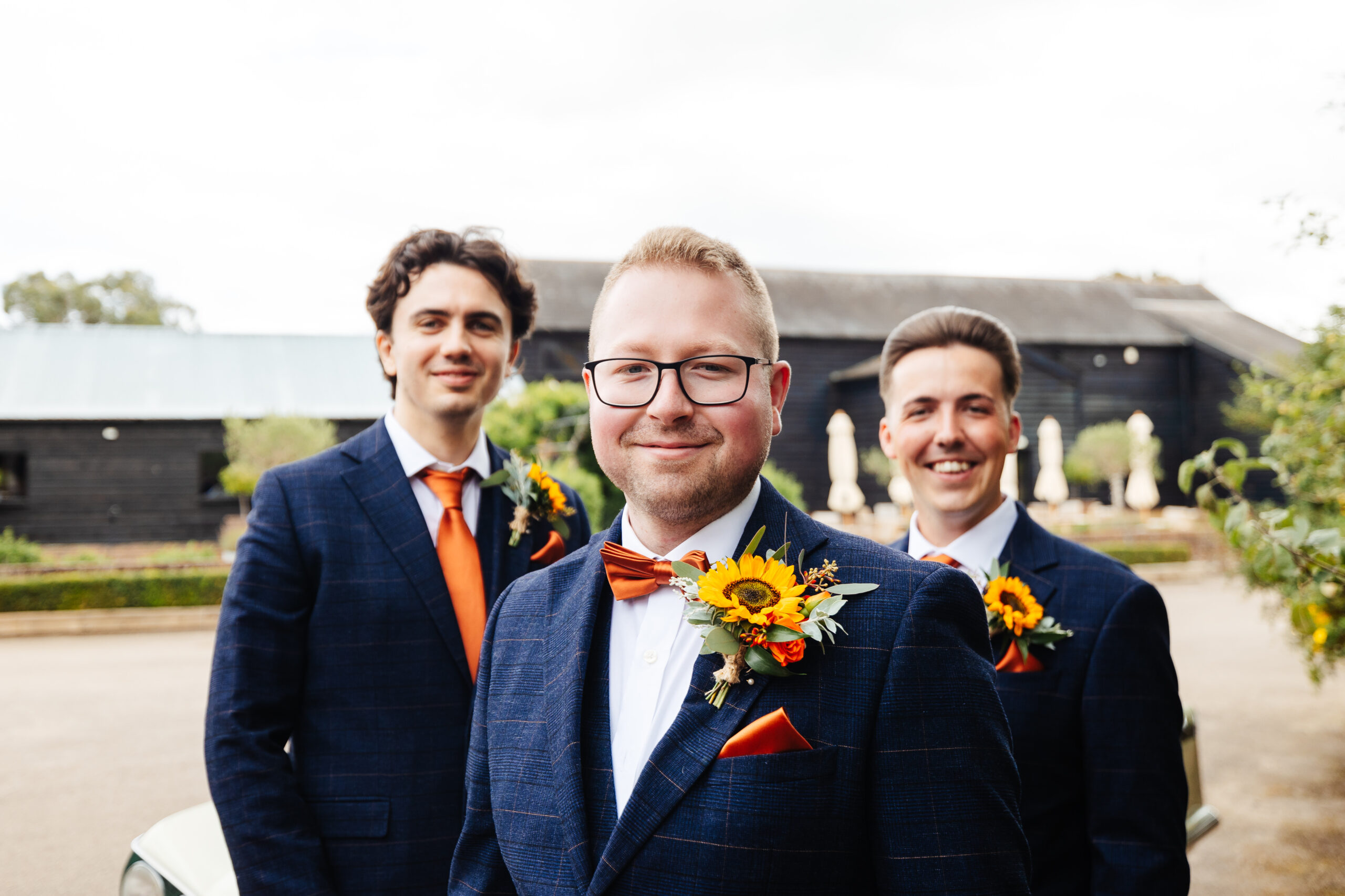 The groom and two men. They are standing outside and you can see the venue in the background. They are wearing navy blue, striped suits with orange ties. The groom has a burnt orange bow tie on. They are looking at the camera and smiling and they have sunflowers as their posies in their top pocket.