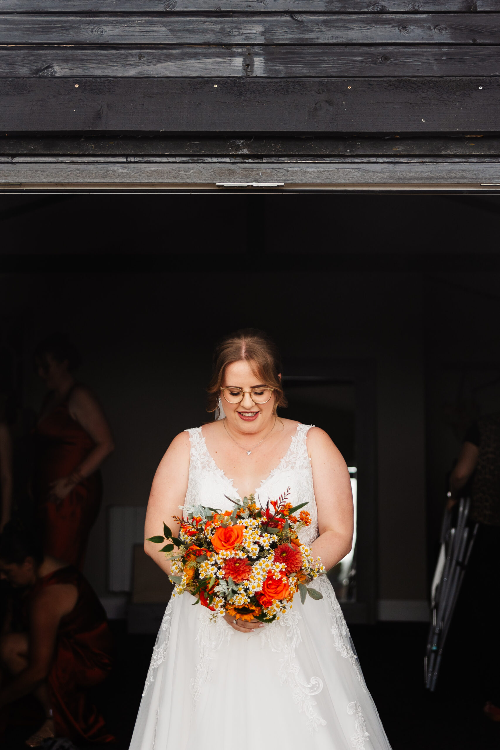 The bride standing in a doorway. She is looking down at a bouquet of bright, orange flowers and smiling.