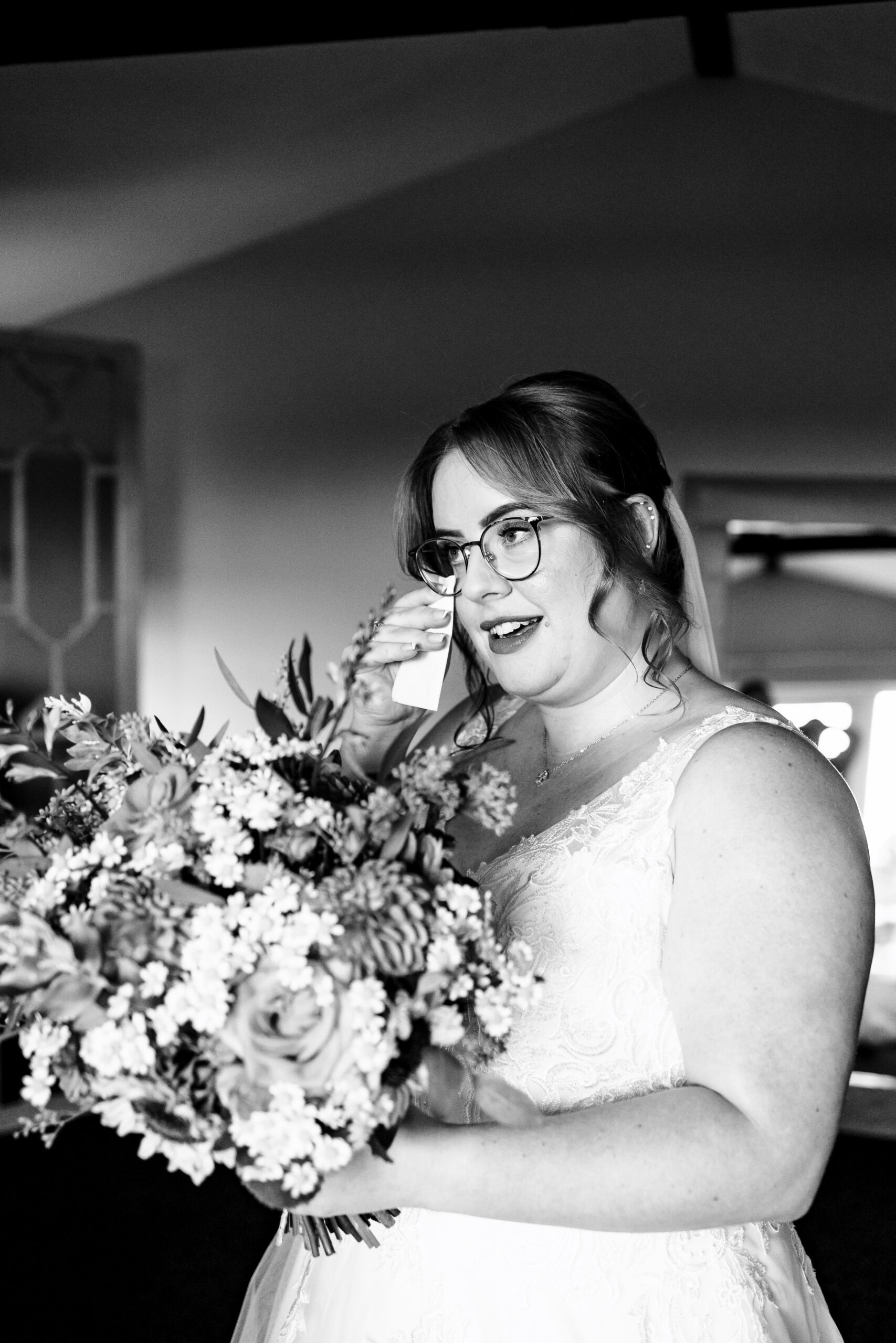 A black and white photo of the bride. She is holding a bouquet of flowers and she has a tissue held up to her face wiping a tear away.