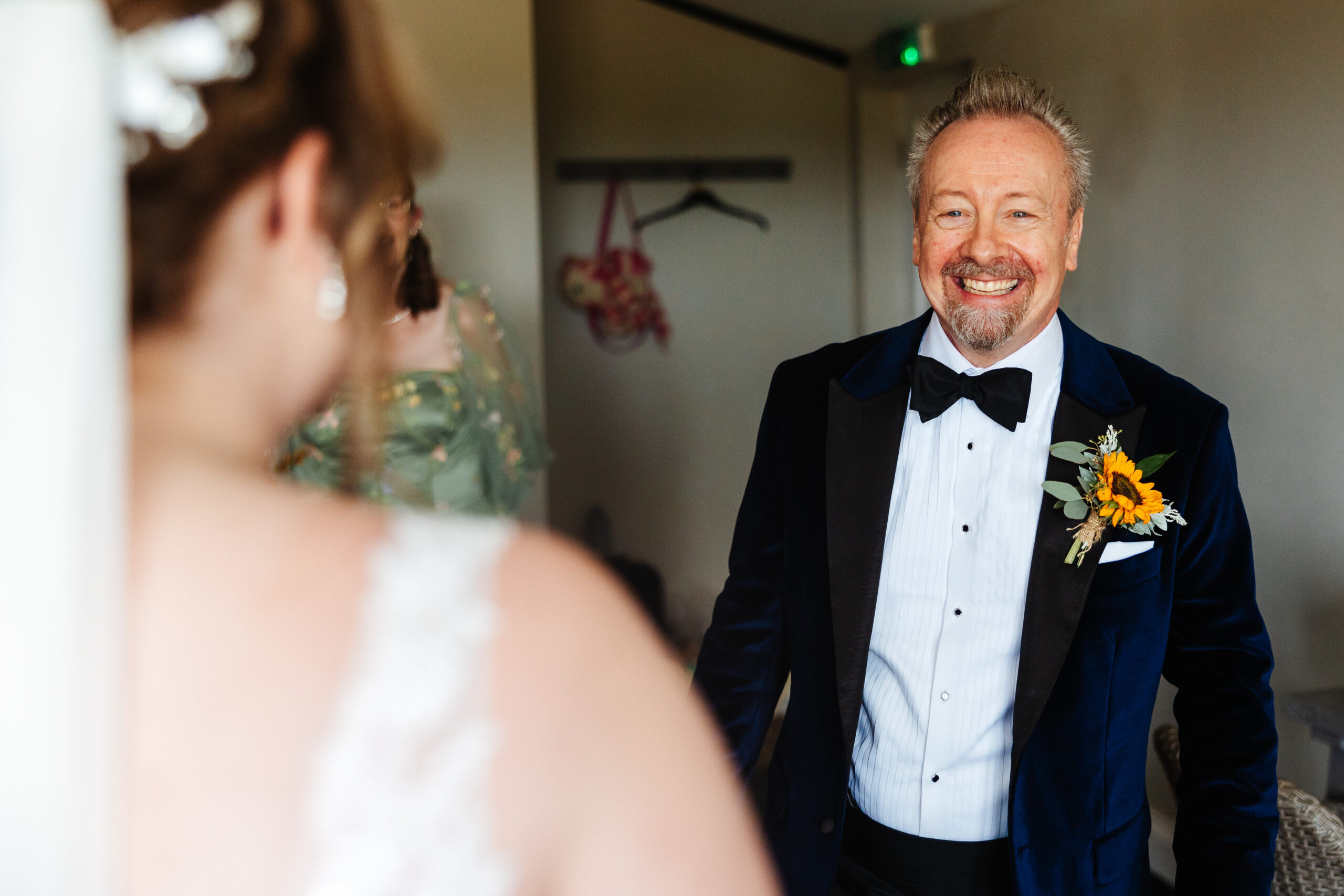 A photo of the father of the bride. He is looking at his daughter in her wedding dress for the first time. He has a huge smile on his face and you can see the shoulder of the bride, blurred in the background. His eyes looks ecstatic and emotional. 