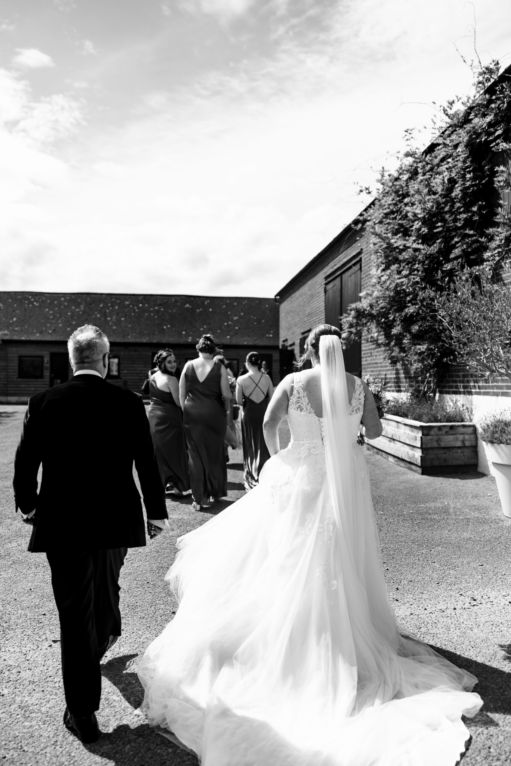 A black and white image of the bride and her father walking across to the venue. You can see her bridesmaids walking in front of her. 