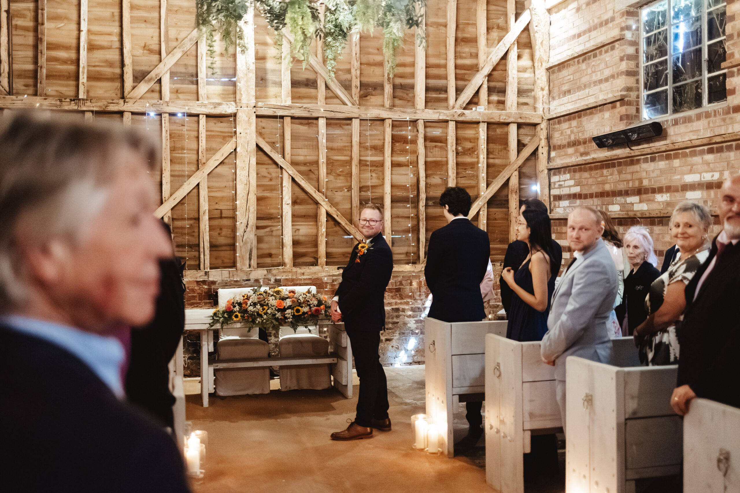 A photo of the groom. He is stood at the top of the alter, looking over his shoulder and smiling. He is looking at his bride. You can see other guests looking at her too and smiling.