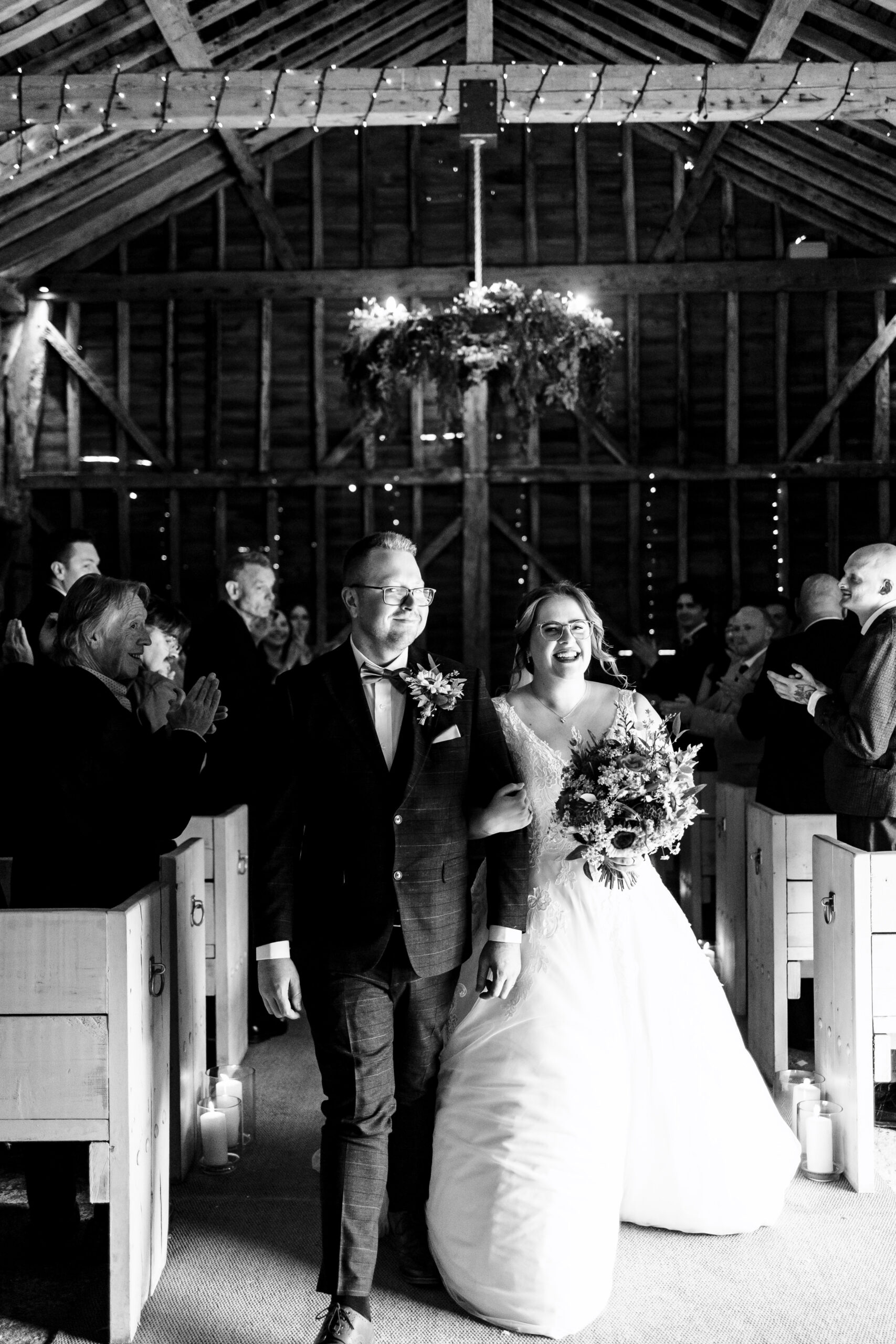 A black and white image of the newlyweds walking out as husband and wife. They are smiling and walking towards the camera. There are guests in the background who are clapping.