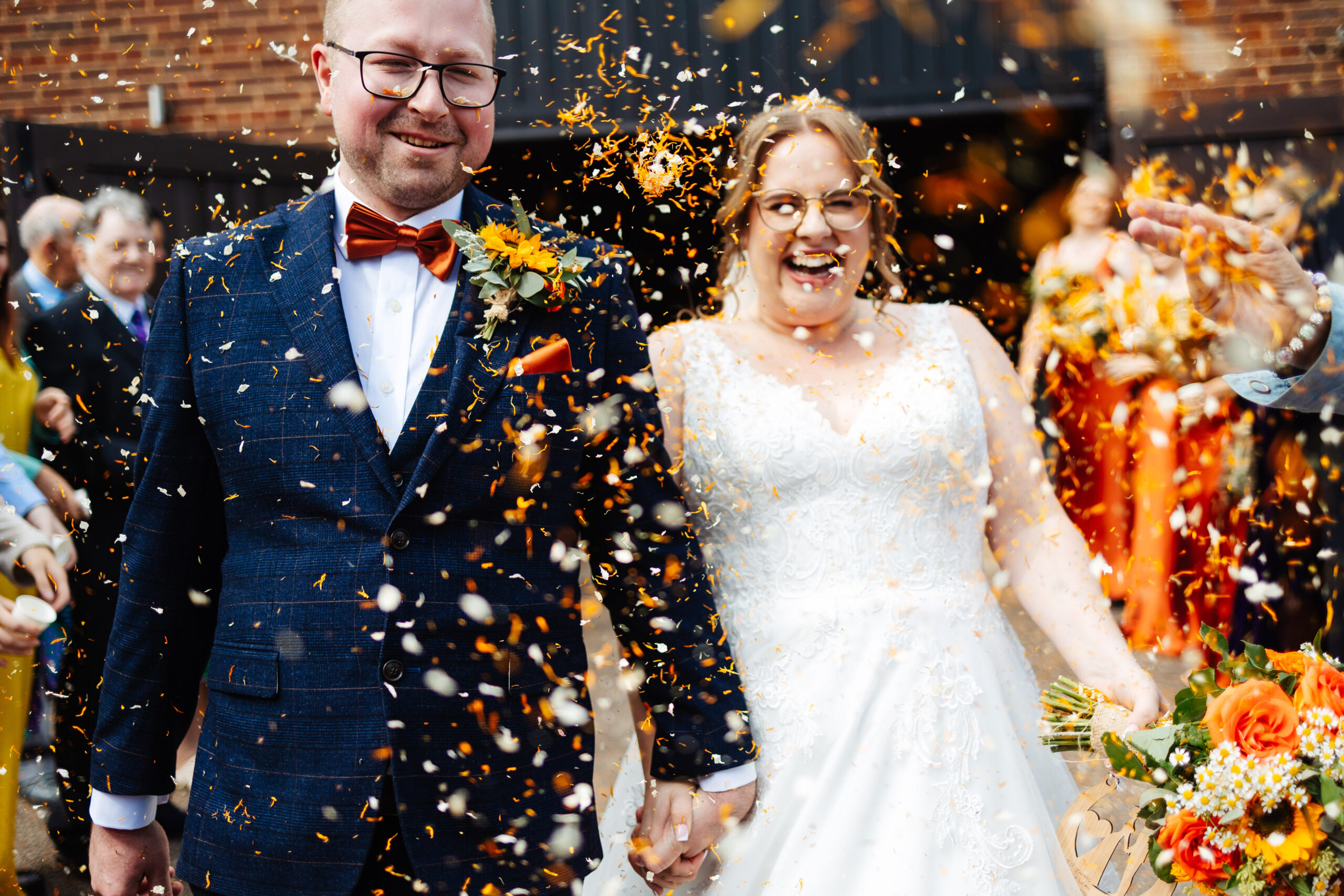 The bride and groom during their confetti shot. They are smiling and there is orange confetti all around them. You can see guests throwing it in the air towards them.