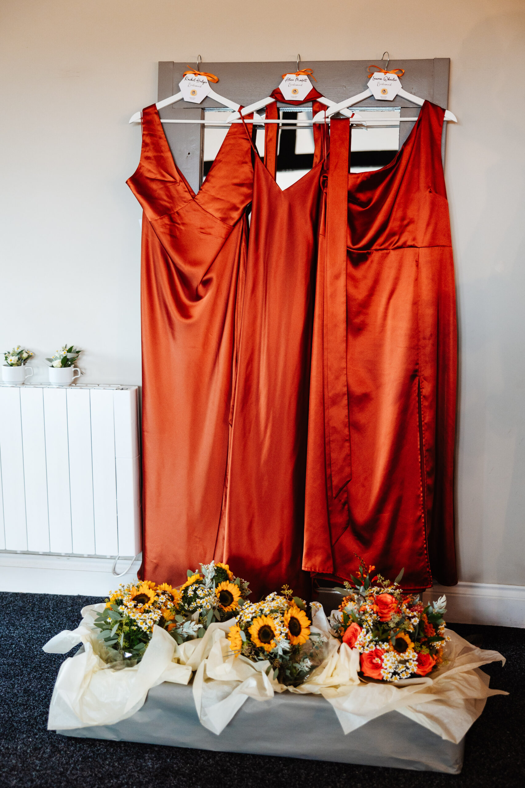 Three, burnt orange dresses hanging up. They have bouquets of flowers at the bottom of them which contain bright yellow sunflowers.