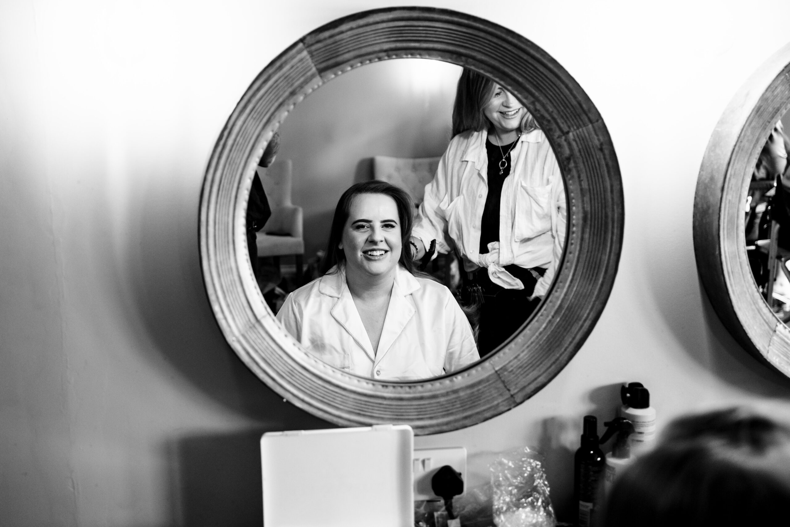 A black and white image of the bride in her silk pyjamas. She is having her hair done and you can see the hairdresser. The image is a reflection in the mirror and the bride is smiling.