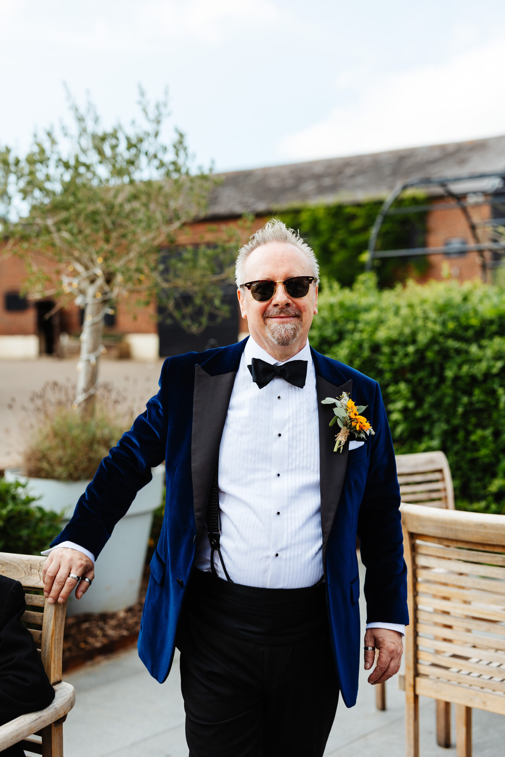 The father of the bride. He is wearing sunglasses and is looking at the camera and smiling. He is wearing a suede navy jacket with a white shirt and a black bow tie. 