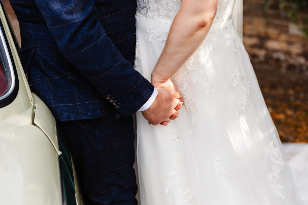 The bride and grooms hands. They are holding hands and you can see the suit and dress in the background.