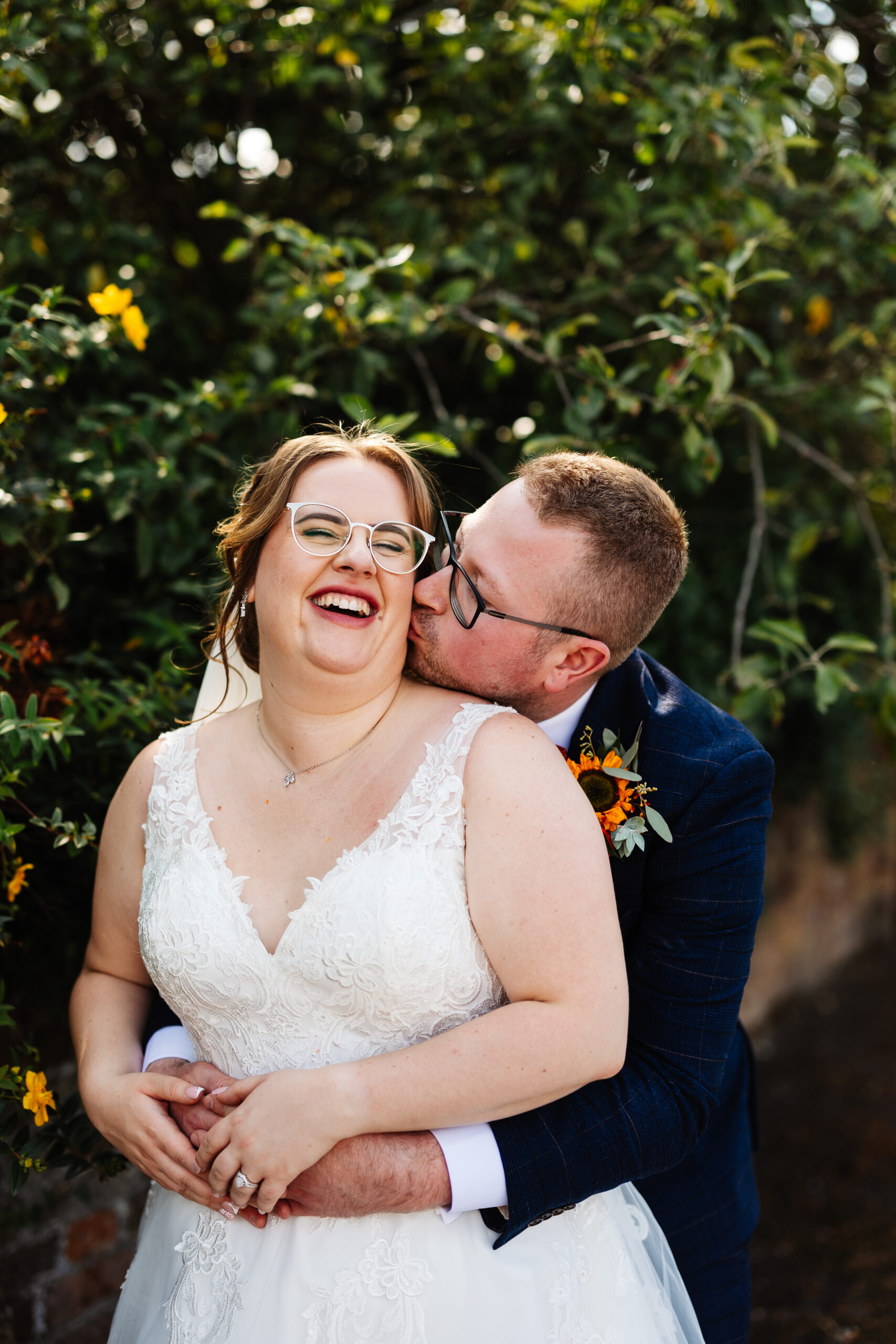 The bride and groom outside in front of some greenery. The groom is behind the bride and is kissing her on the cheek. She has her eyes closed and she is smiling.