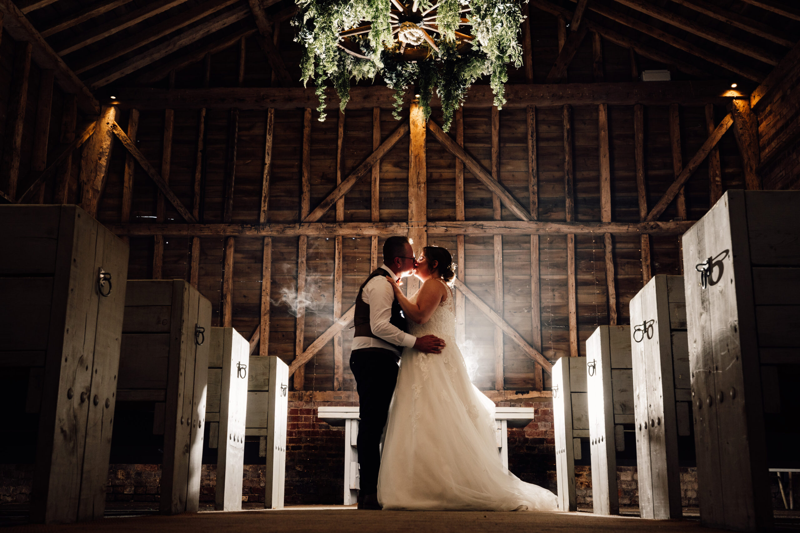 A photo of the bride and groom in a barn. They are kissing and the sunlight is pouring in through a gap in the wood.