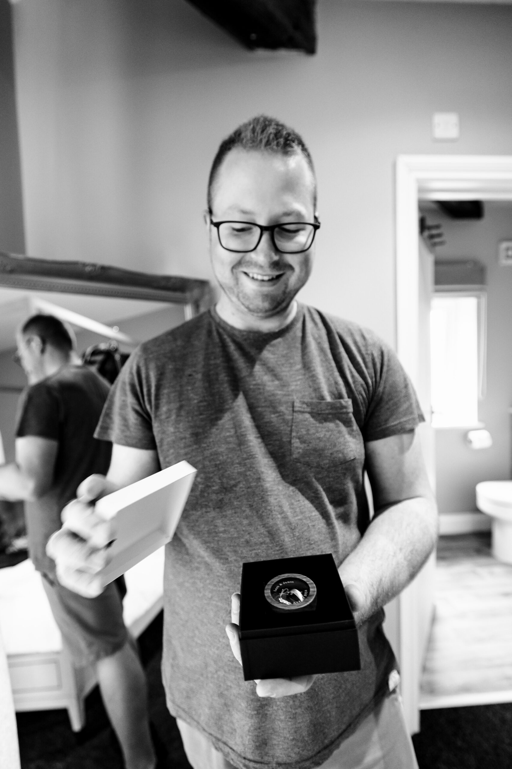 A black and white image of the groom. He is in casual clothes, a t-shirt and trousers. He is looking down at a box containing the ring box. He is smiling.