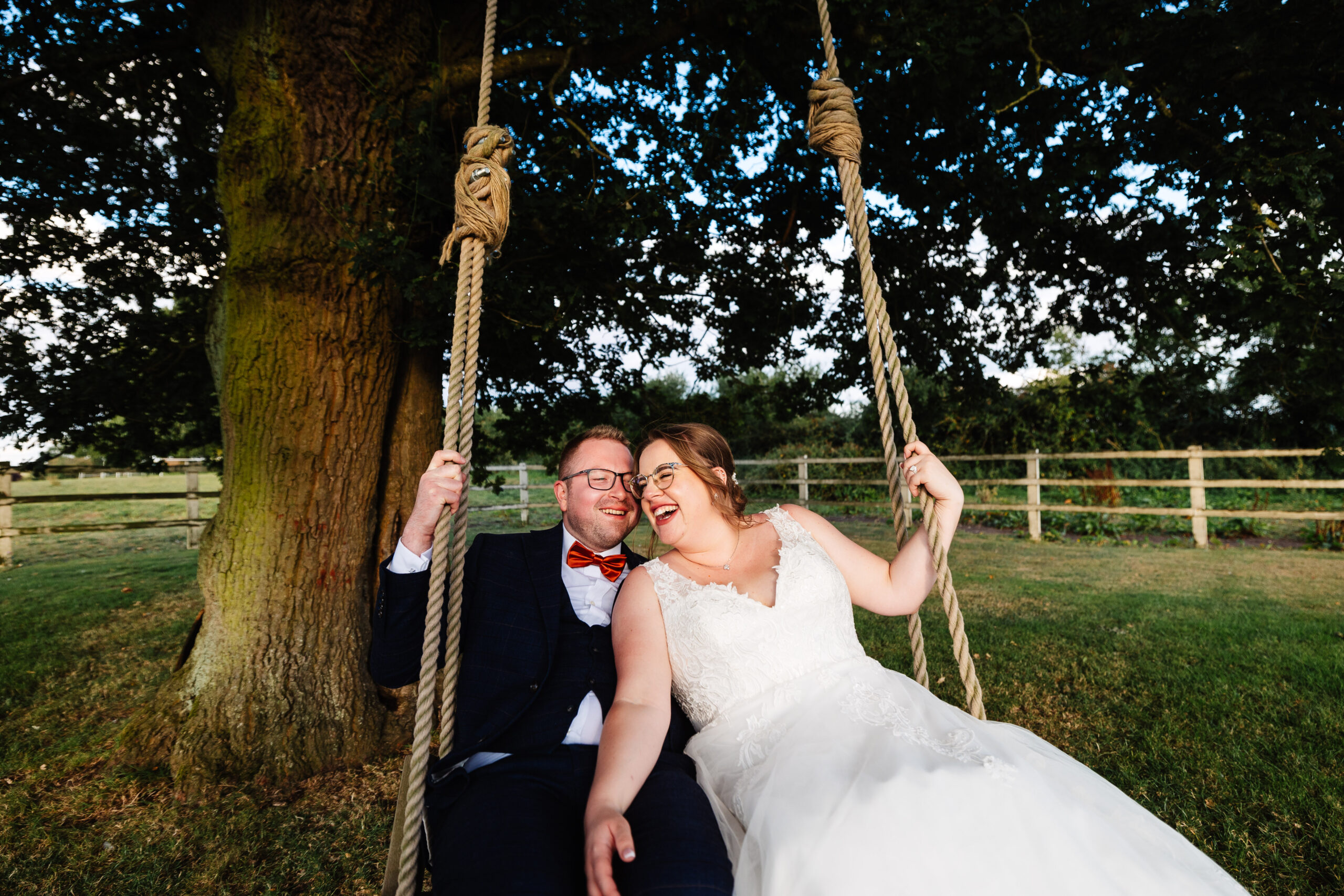 The bride and groom on an outdoor, tree swing. They are close to each other and smiling.