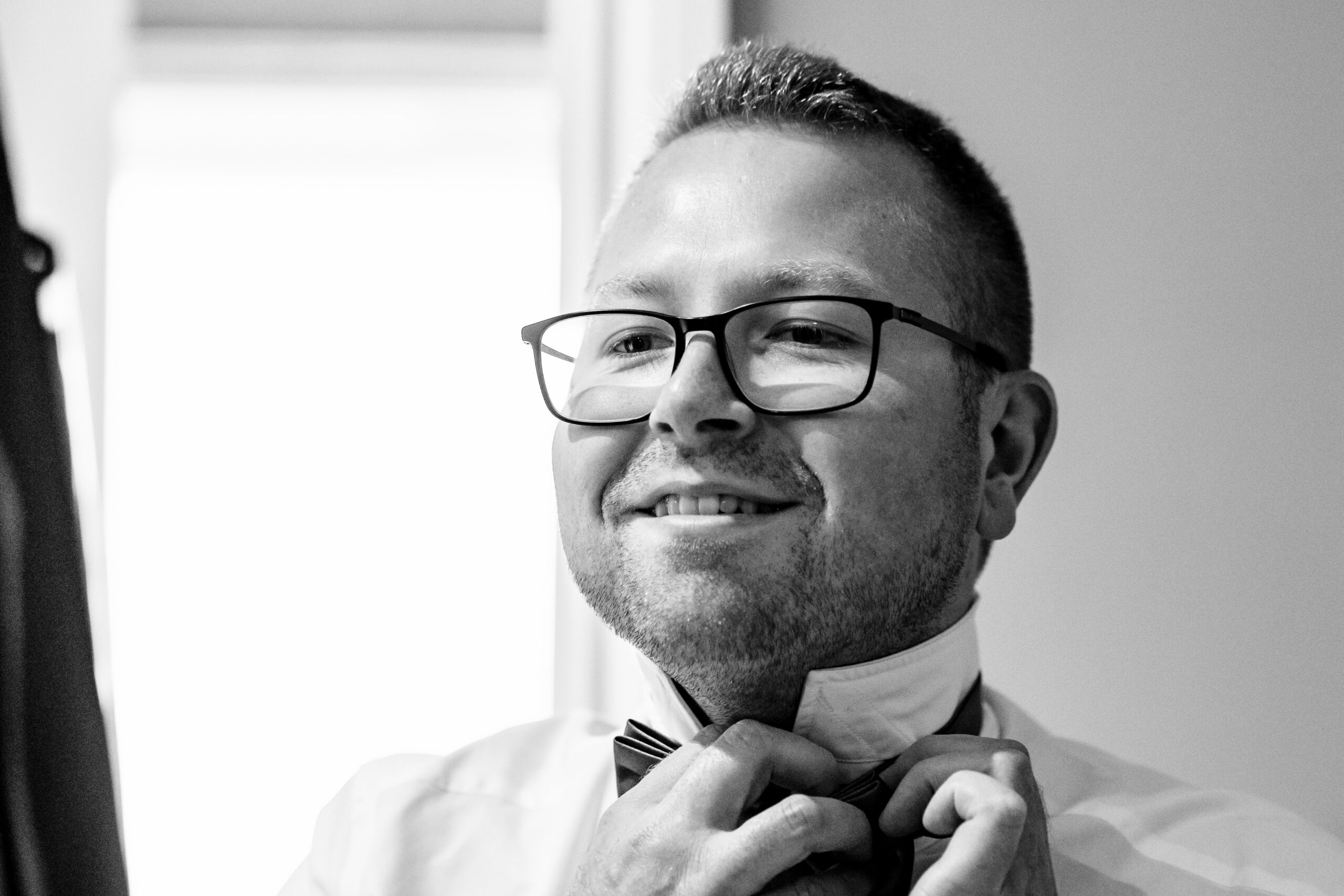 A black and white image of the groom. He is looking in the mirror and he is smiling. He is wearing a shirt and is adjusting his bow tie.