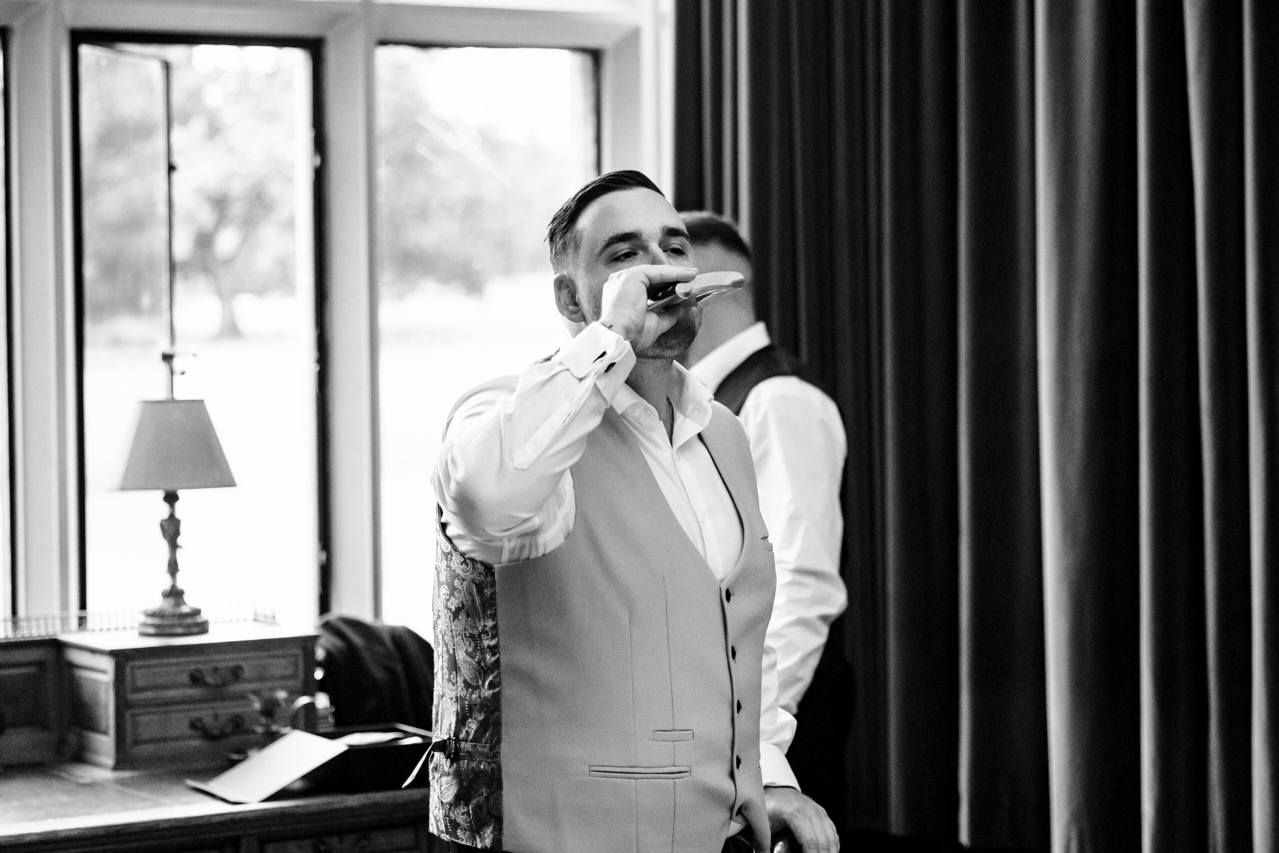 A black and white photo of the groom in his shirt and waistcoat and drinking from a hip flask.