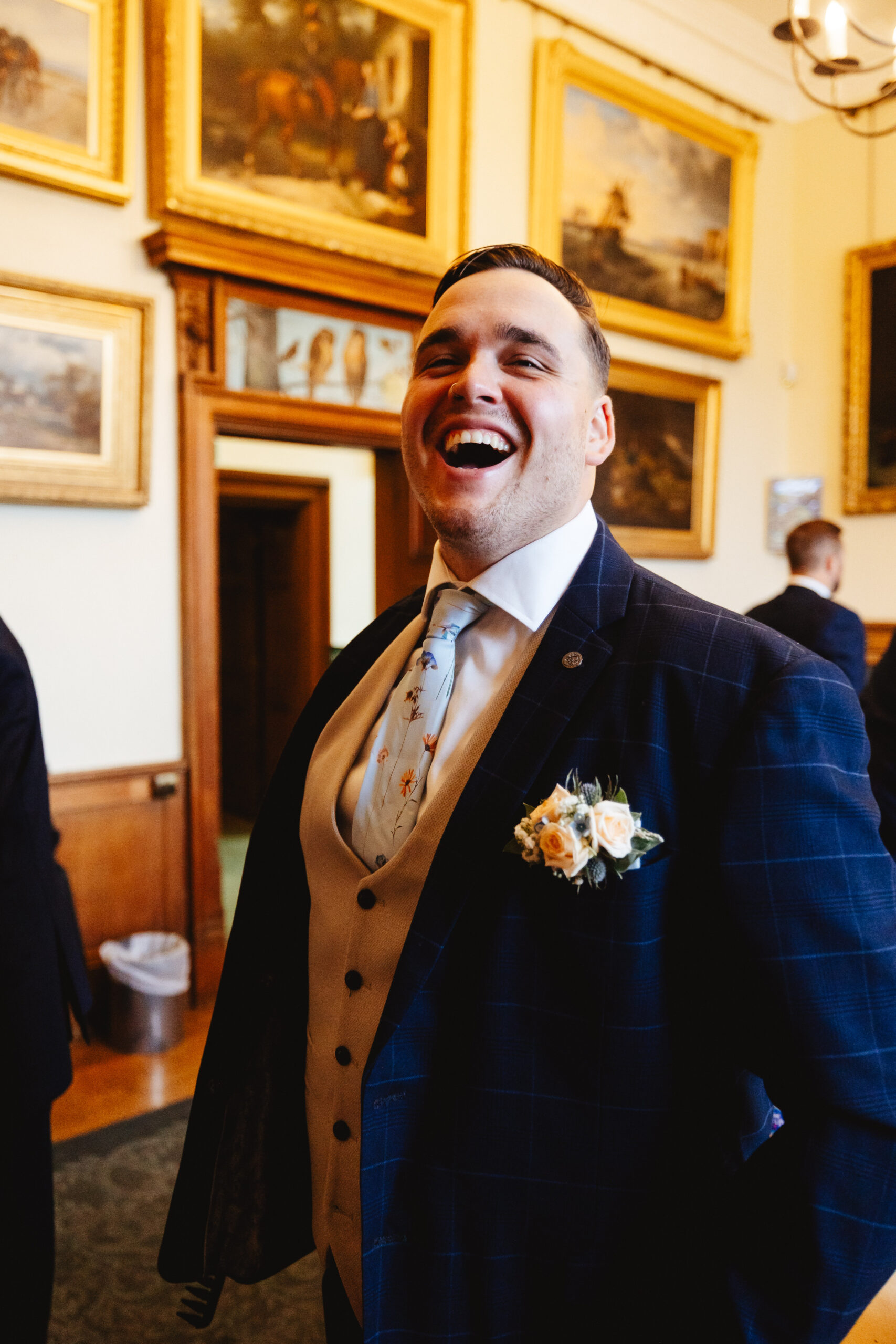 The groom. He is dressed in a pin striped navy blue suit. He has a pale blue tie on with several colourful flowers on. He is smiling at the camera.