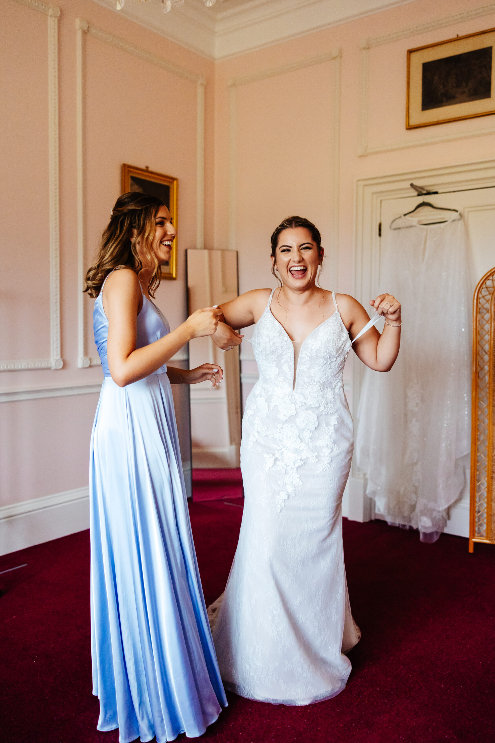 The bride getting in to her wedding dress. She is wearing it but just waiting on her left strap to be put on her arm. Her bridesmaid is helping he and she is wearing a lilac dress.