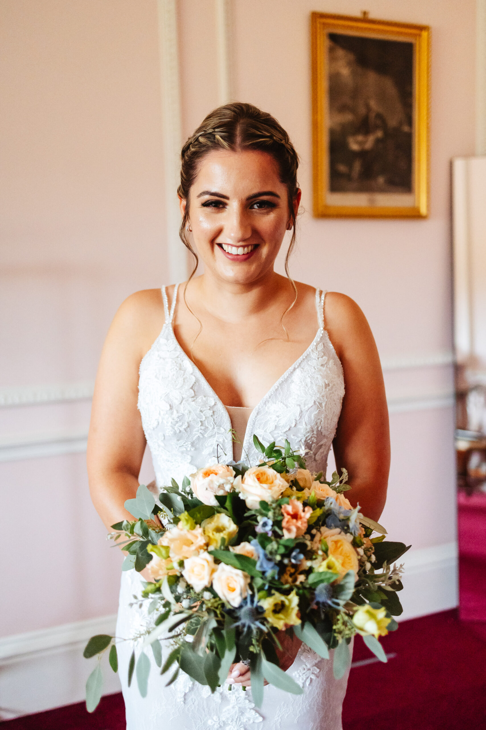 The bride smiling at the camera. She is looking at the camera looking excited. She is wearing her wedding dress and has a bouquet of flowers in front of her. They are a mixture of pale orange, yellow and blue.