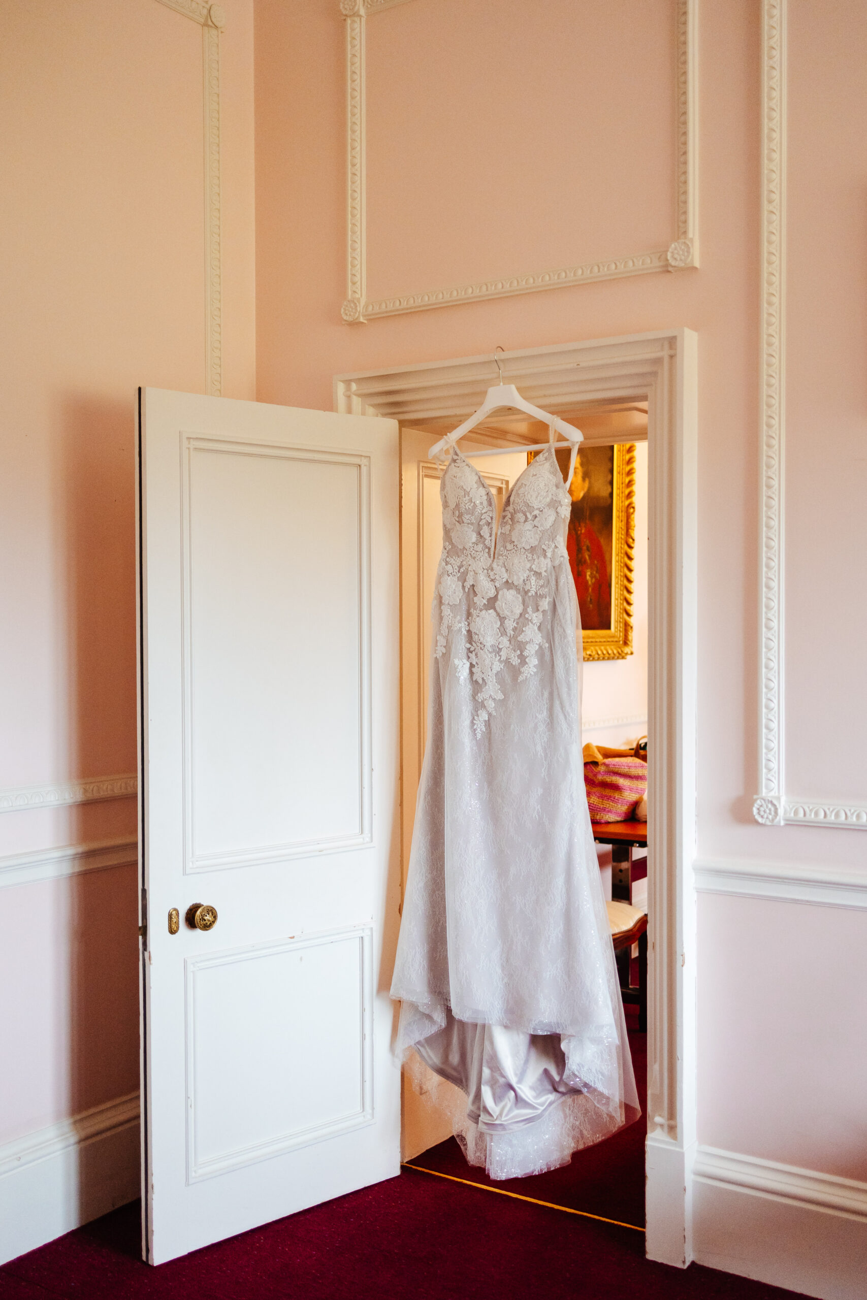 A photo of the bride's dress hanging in the doorway. It is a long dress with a beaded and lace detailed front.