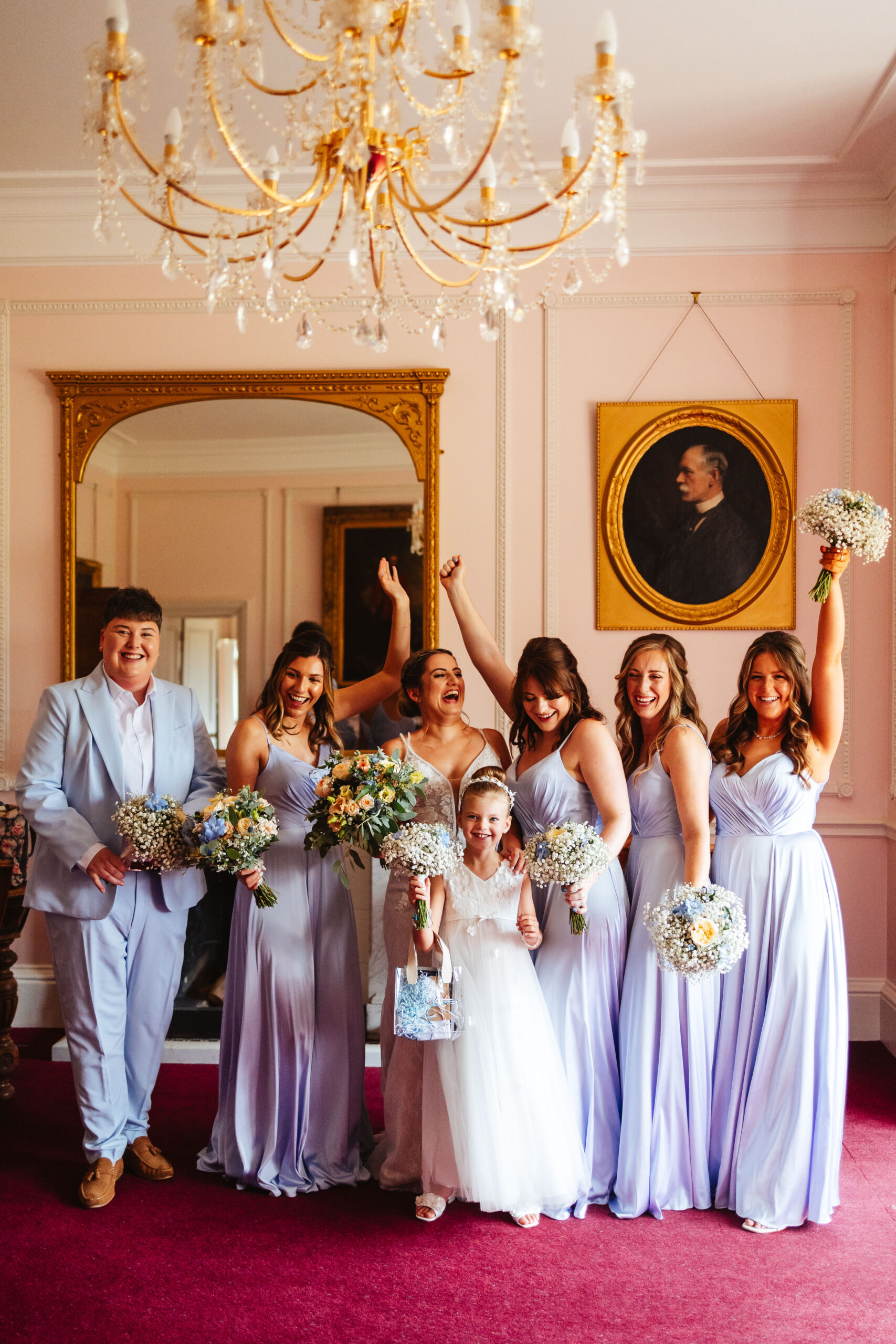 The bride and her bridesmaids. The bridesmaids are wearing long, lilac dresses and one of them is in a matching lilac suit. They are all happy and have their arms and flowers in the air.