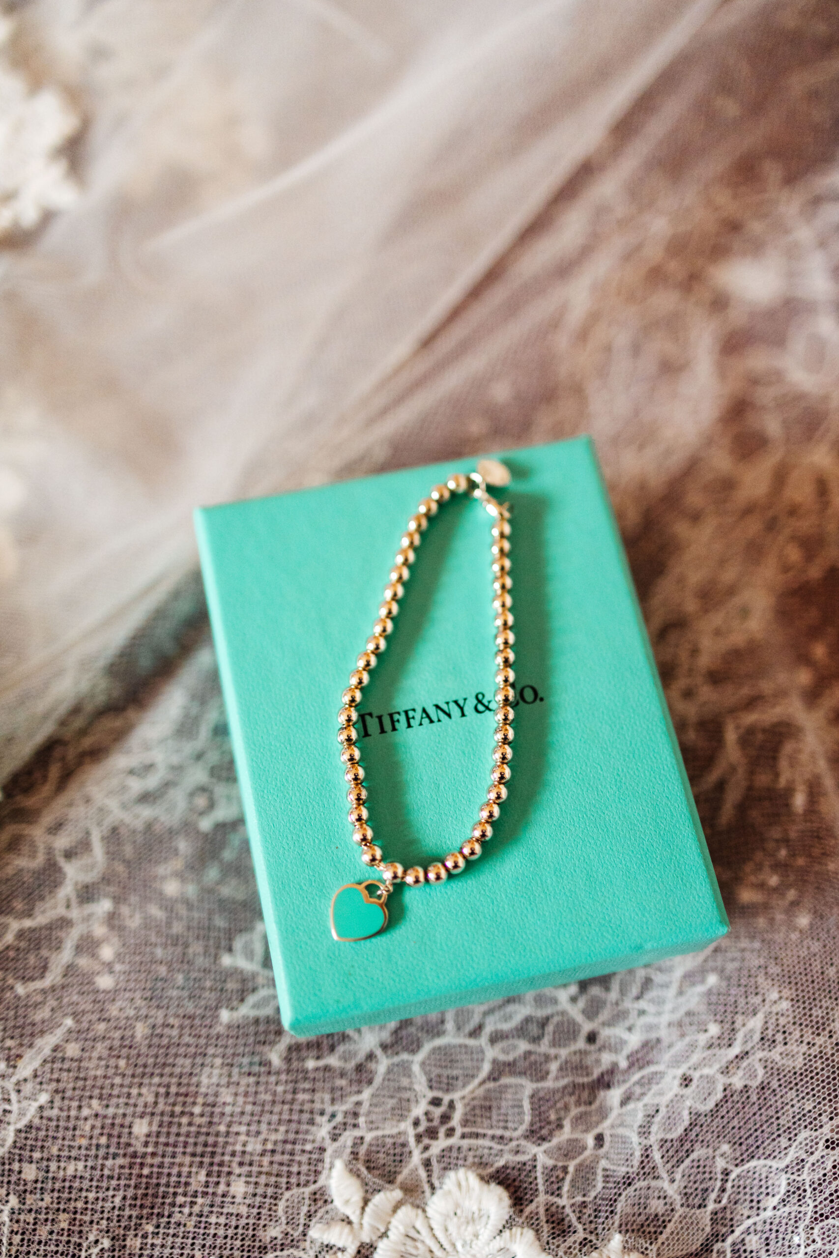 A gold beaded bracelet with a turqouise heart charm on it. It is on a Tiffany & Co. box.