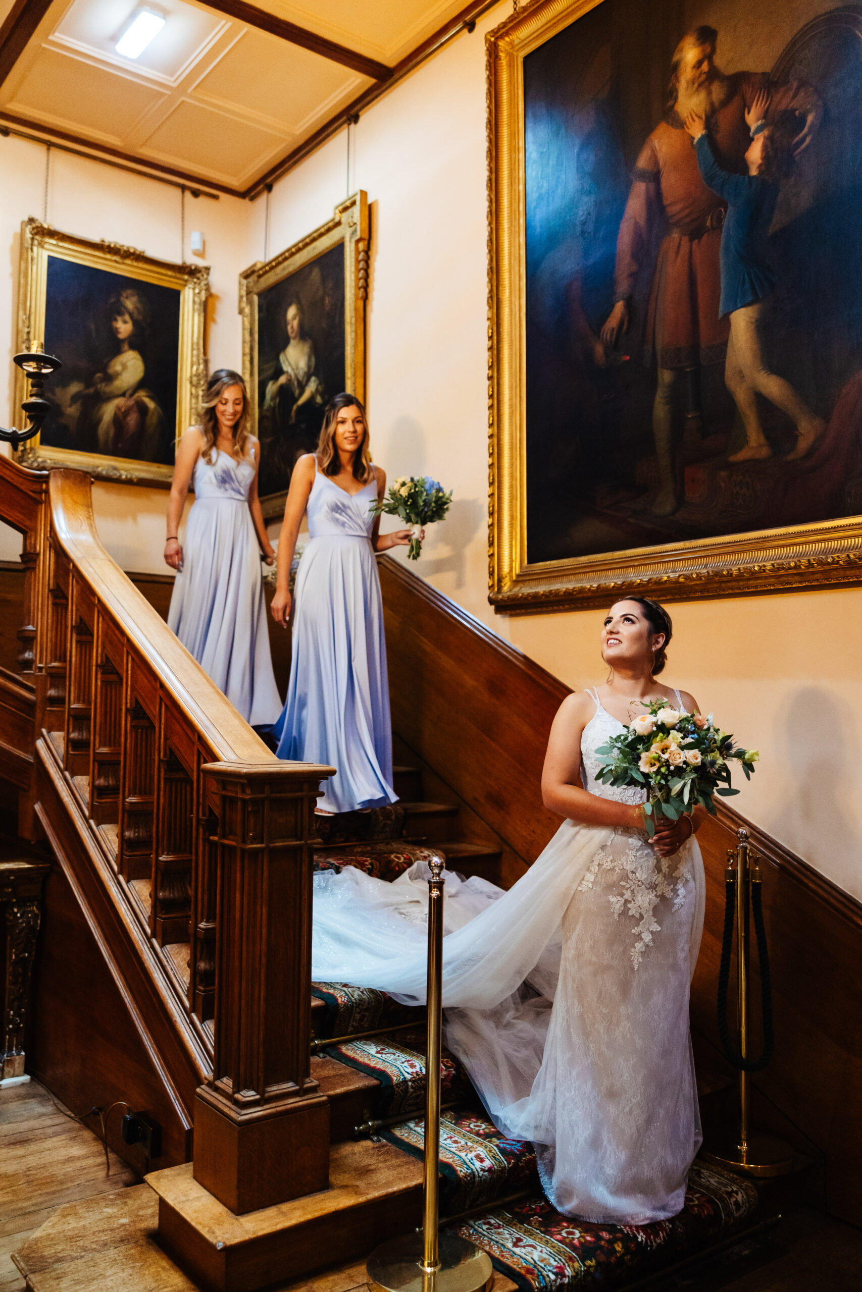 The bride walking down an old, wooden staircase. Her bridesmaids are behind her. They are all smiling.