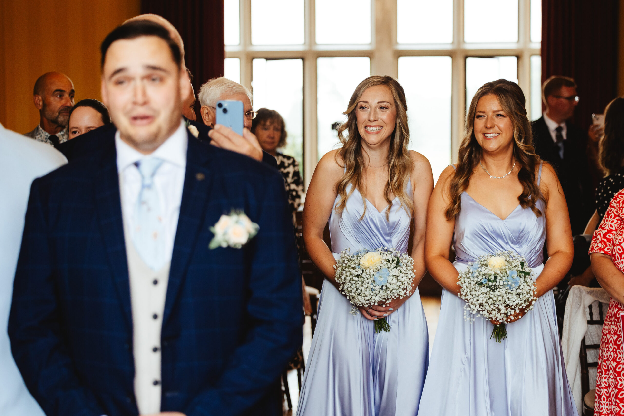 The groom at the top of the alter. Two bridesmaids are walking behind him. He looks emotional. 