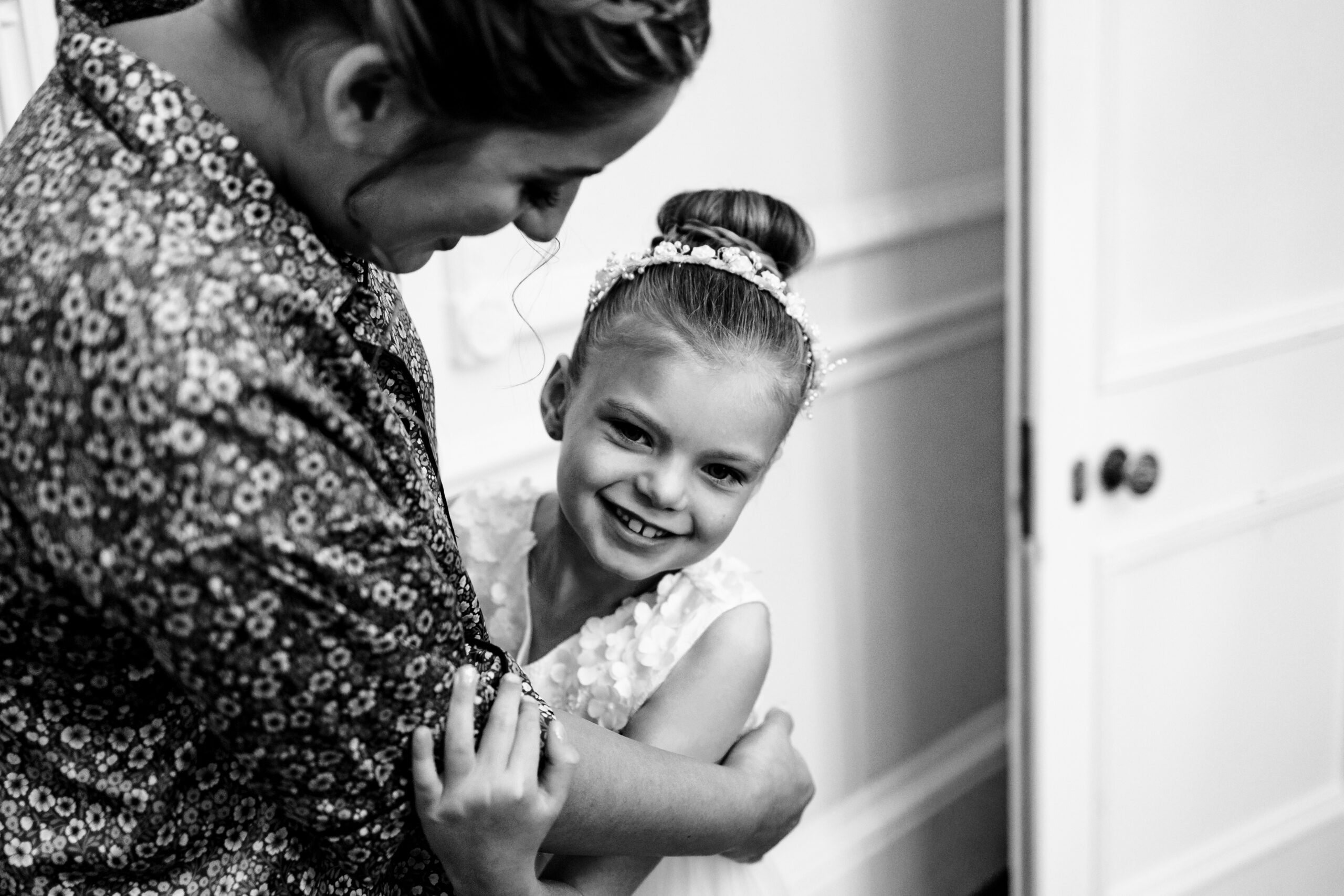 A black and white photo of a little girl in a white dress. She has her hair up in a bun and has a headband on too. She is hugging the bride and is smiling.