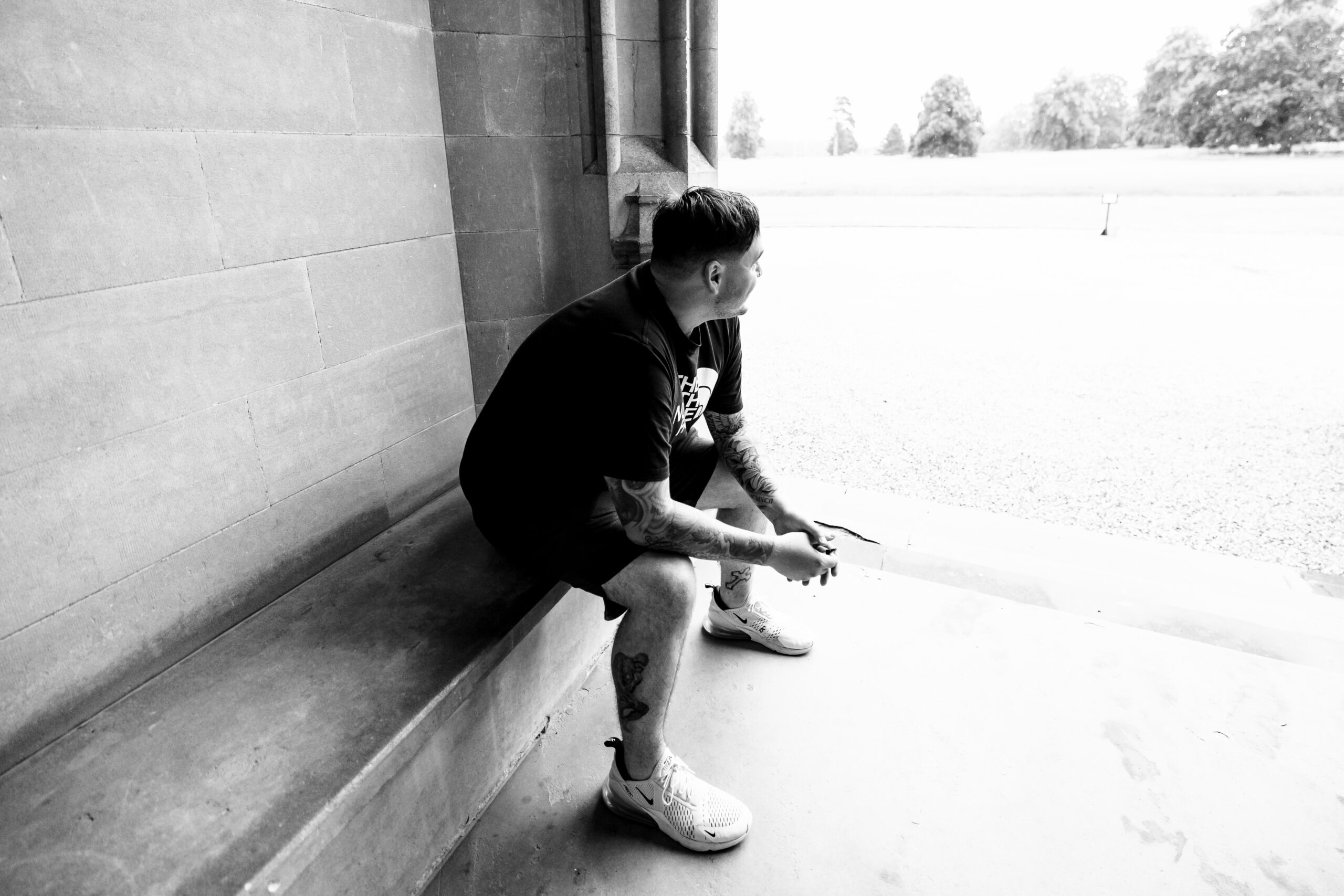 A black and white image of the groom sat down looking out in to the distance. He is sat on a bench of his wedding venue and is smiling.