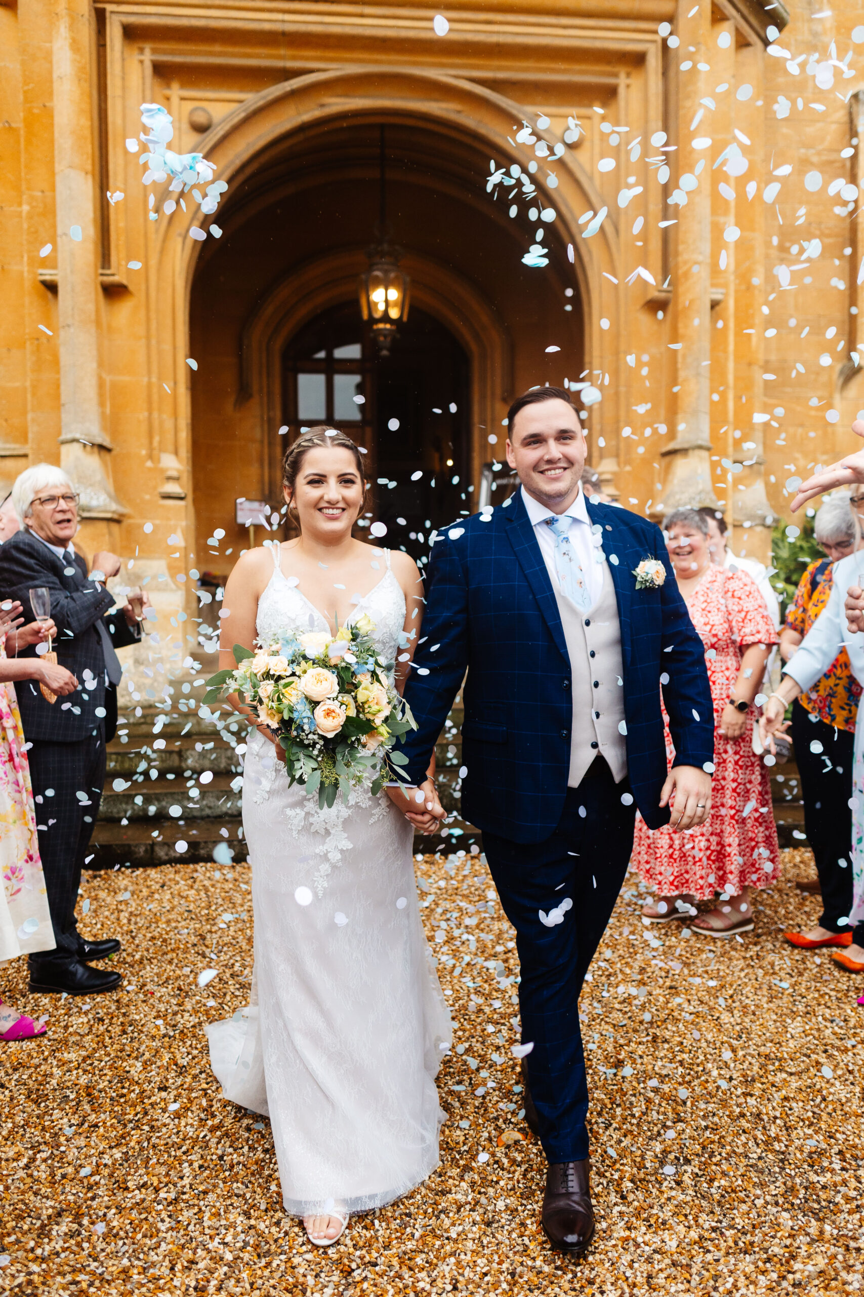 The bride and groom outside. They are surrounded by their guests whoa are throwing pale blue confetti in to the air. The bride and groom are smiling and holding hands.