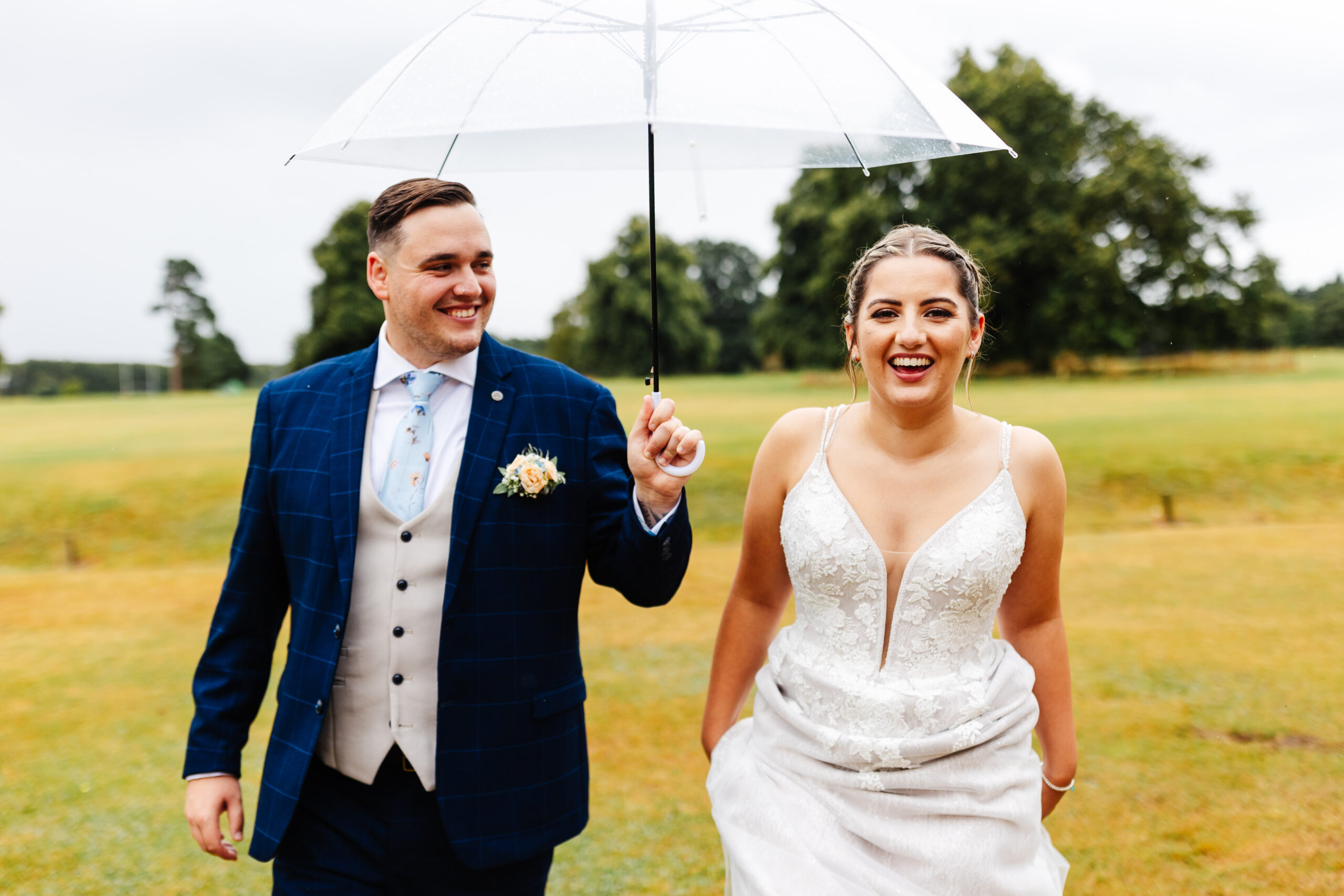 The bride and groom outside. The bride is looking at the camera and the groom is looking at her. He is holding an umbrella over them.