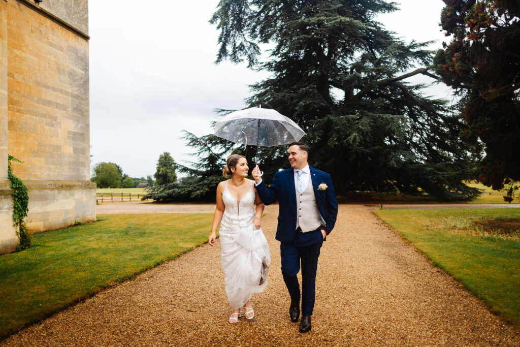 bride and groom walking alongside shuttleworth house with umbrella, bride is holding her dress off the floor and laughing