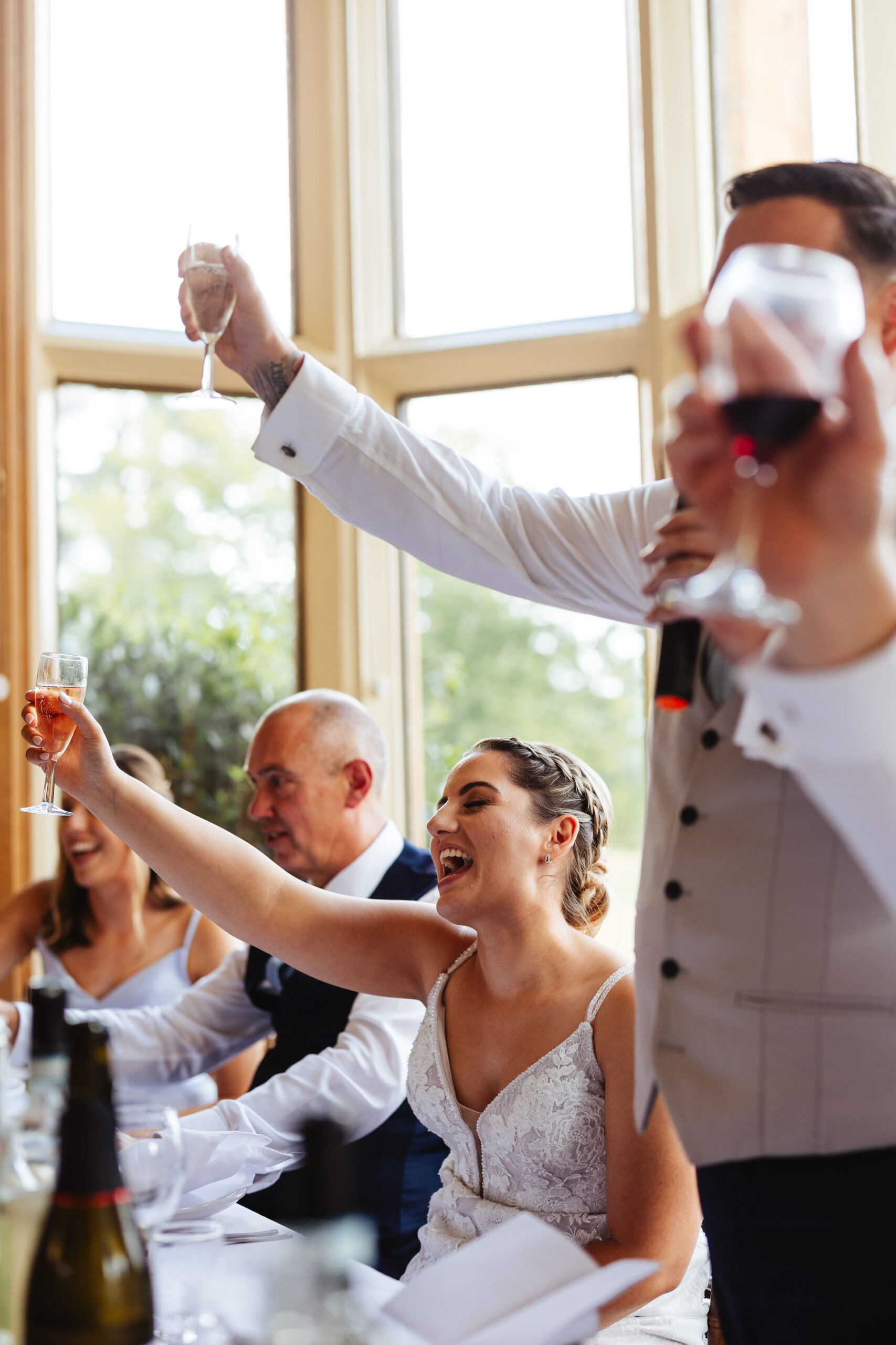 The bride is sitting down and is holding a glass of prosecco in her hand in the air and smiling. Her groom is stood up holding a microphone in one hand and is cheersing with the other. 