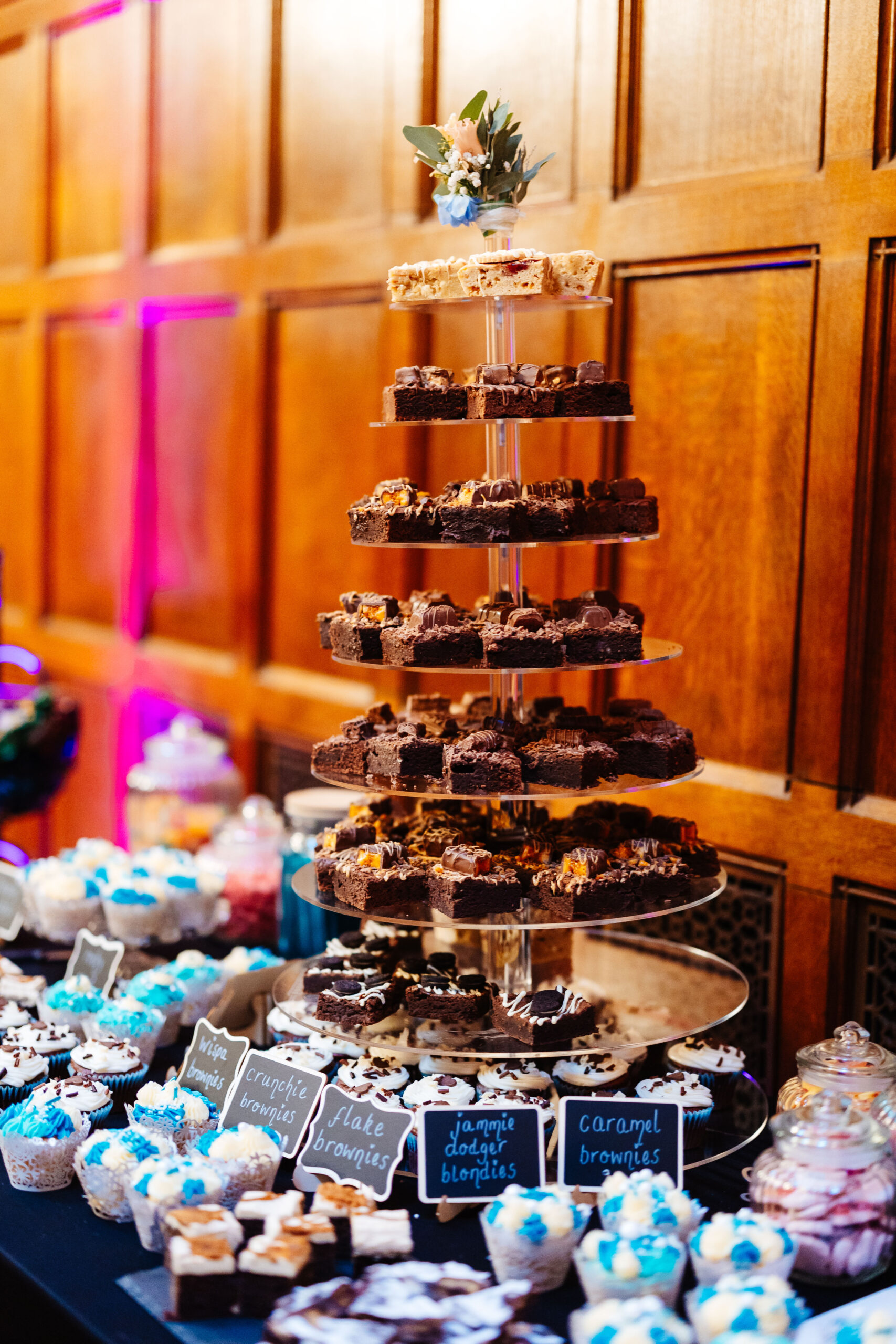 An eight tiered stand with lots of chocolate cakes on. This is surrounded by mini cupcakes in all flavours such as caramel brownie, crunchie brownie, jammie dodger blondies